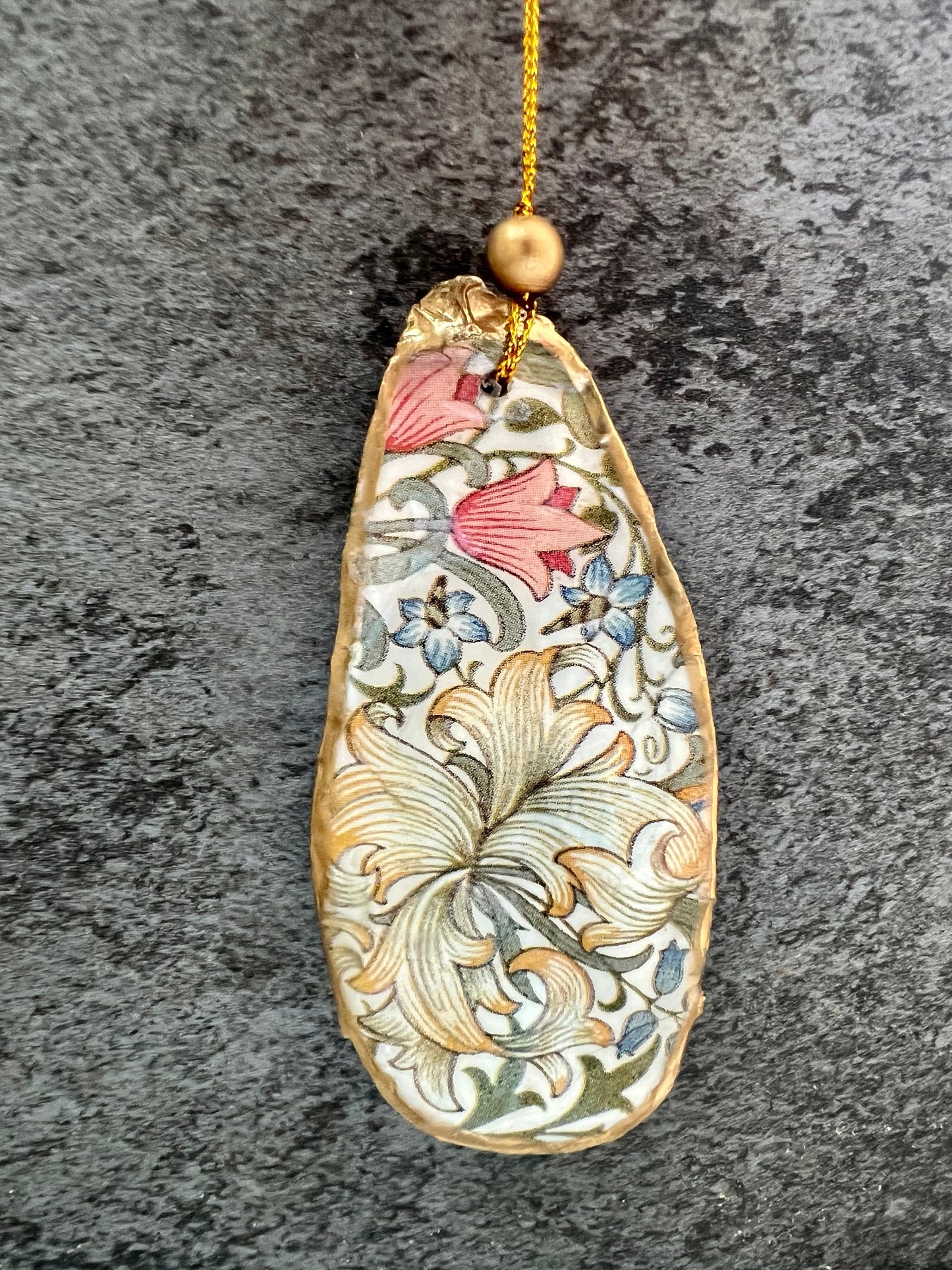 Handmade Dorset oyster shell hanging ornaments featuring William Morris inspired Golden Lily and Granville floral designs in soft green, blush pink and gold tones. Natural oyster shells finished with liquid gold leaf edging and sealed in glossy resin. Decorative hanging ornaments with gold cord and bead detail, suitable for luxury Christmas décor, coastal interiors, cottagecore styling or elegant year-round home decoration.