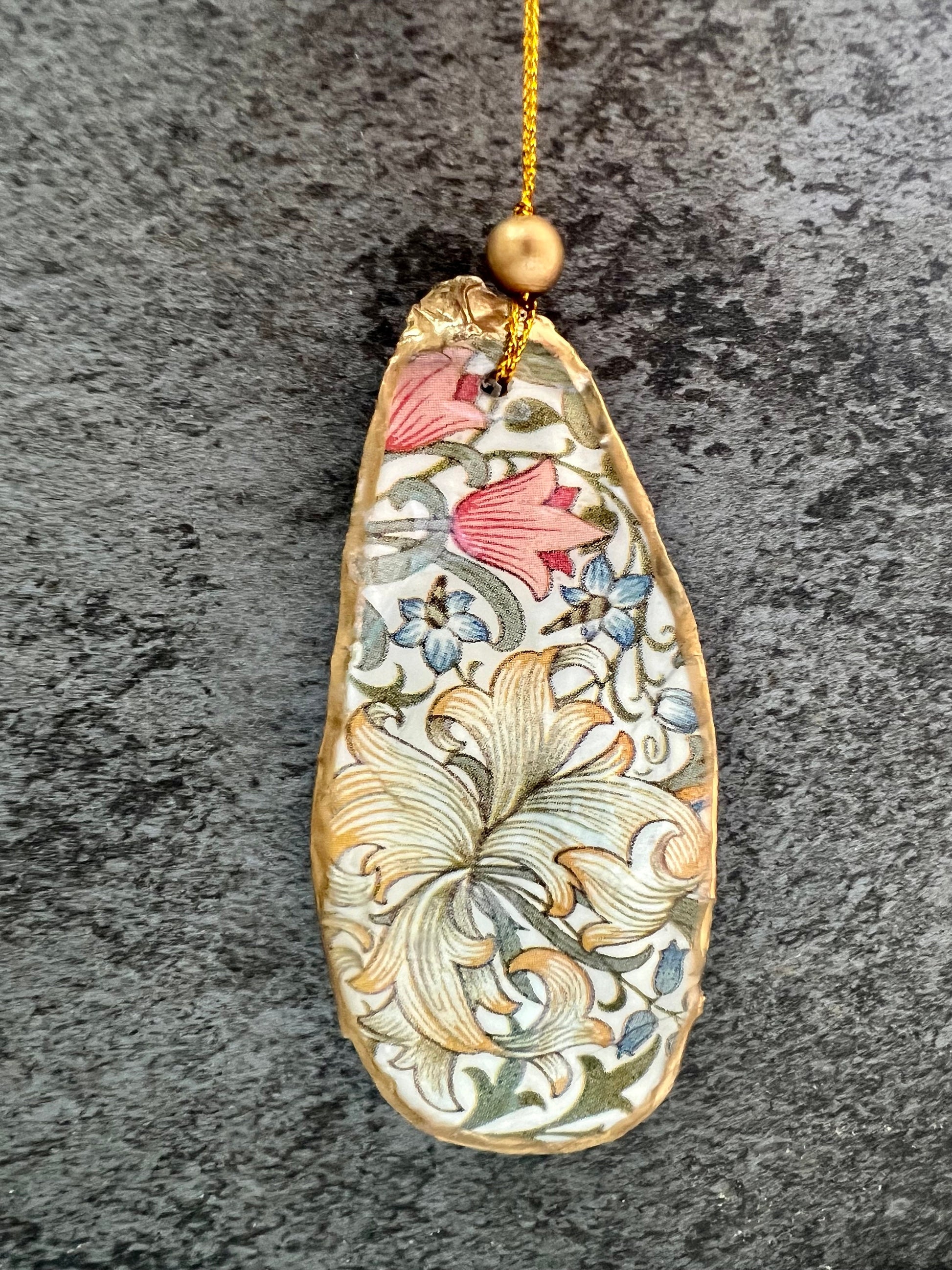 Handmade Dorset oyster shell hanging ornaments featuring William Morris inspired Golden Lily and Granville floral designs in soft green, blush pink and gold tones. Natural oyster shells finished with liquid gold leaf edging and sealed in glossy resin. Decorative hanging ornaments with gold cord and bead detail, suitable for luxury Christmas décor, coastal interiors, cottagecore styling or elegant year-round home decoration.