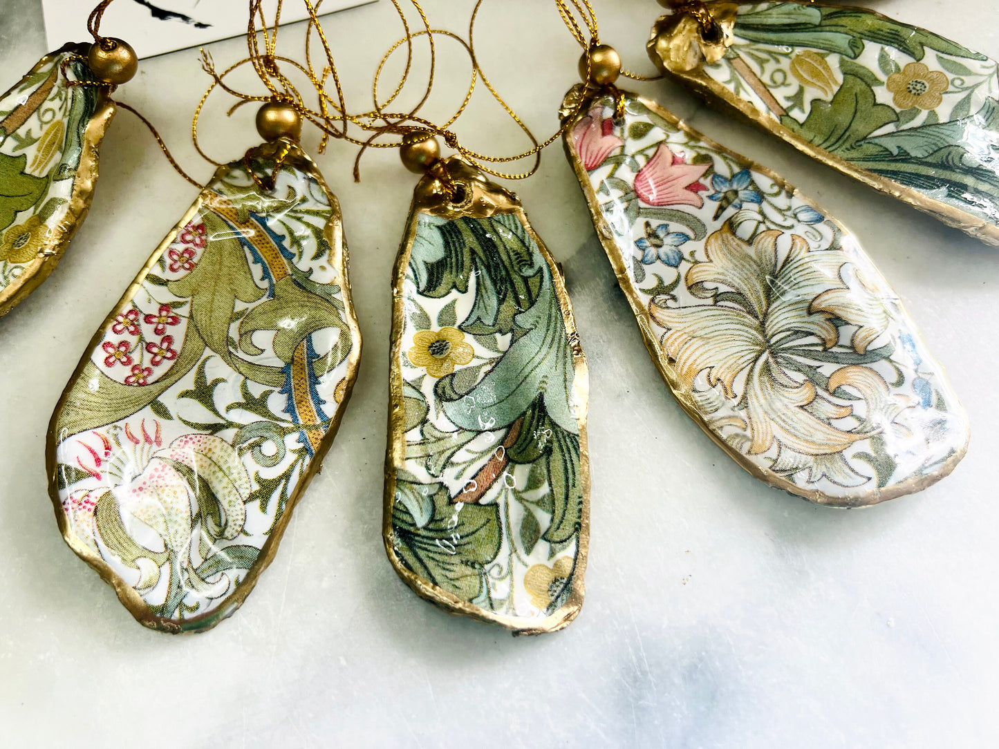 Handmade Dorset oyster shell hanging ornaments featuring William Morris inspired Golden Lily and Granville floral designs in soft green, blush pink and gold tones. Natural oyster shells finished with liquid gold leaf edging and sealed in glossy resin. Decorative hanging ornaments with gold cord and bead detail, suitable for luxury Christmas décor, coastal interiors, cottagecore styling or elegant year-round home decoration.