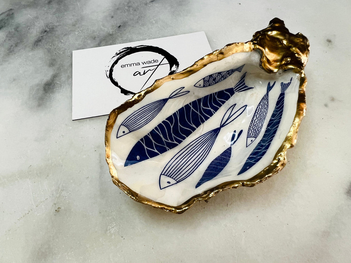 Hand-gilded Dorset oyster shell trinket dish featuring a blue fish motif, edged with liquid gold leaf and sealed in high gloss resin. Coastal ring holder and nautical home decor piece displayed with multiple shells. Sustainable oyster shell art handmade in Surrey, ideal as a jewellery dish, wedding favour, beach house accessory or ocean lover gift.