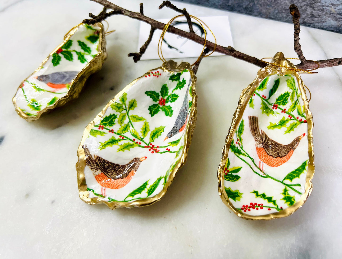Handmade Dorset oyster shell Christmas ornament featuring red and orange winter birds perched amongst festive green foliage and berries. Coastal oyster shell trinket dish with hand applied gold leaf edging and glossy resin finish, styled as luxury Christmas décor. Sustainable handcrafted robin-inspired shell decoration available as hanging ornament or jewellery dish, inspired by British winter wildlife and the Dorset coastline.