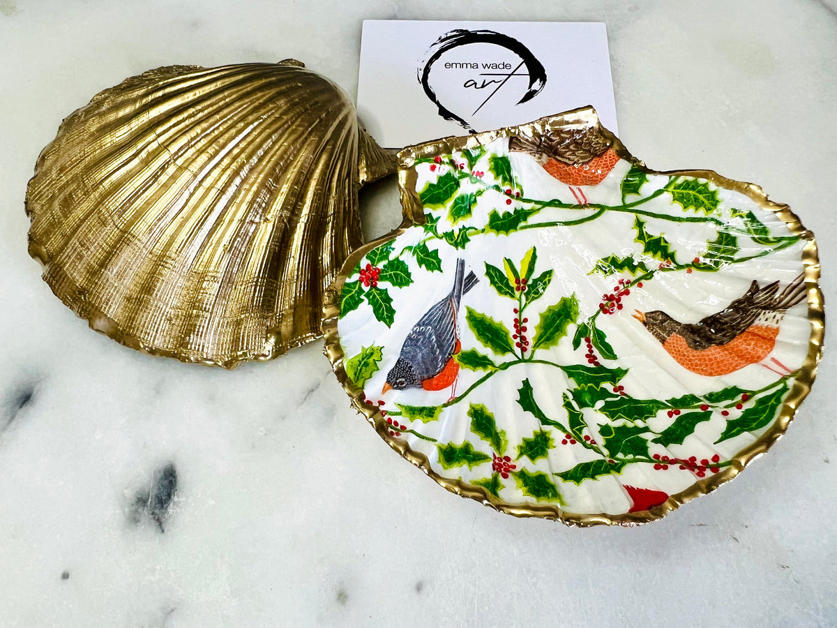 Handmade Dorset scallop shell trinket dish decorated with winter robin and woodland bird illustrations surrounded by festive holly leaves and berries. Natural shell hand finished with liquid gold leaf edging and sealed with glossy resin. Bird décor inspired by coastal cottage interiors and nature styling, suitable as a jewellery dish, ring tray, festive ornament, robin lover gift, winter home décor or Christmas stocking filler.