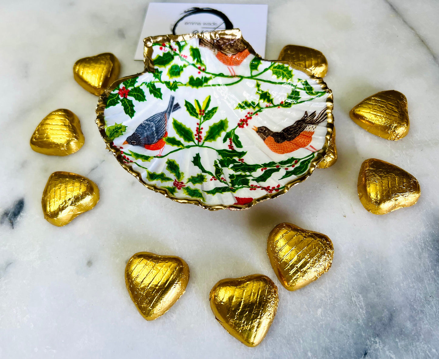 Handmade Dorset scallop shell trinket dish decorated with winter robin and woodland bird illustrations surrounded by festive holly leaves and berries. Natural shell hand finished with liquid gold leaf edging and sealed with glossy resin. Bird décor inspired by coastal cottage interiors and nature styling, suitable as a jewellery dish, ring tray, festive ornament, robin lover gift, winter home décor or Christmas stocking filler.