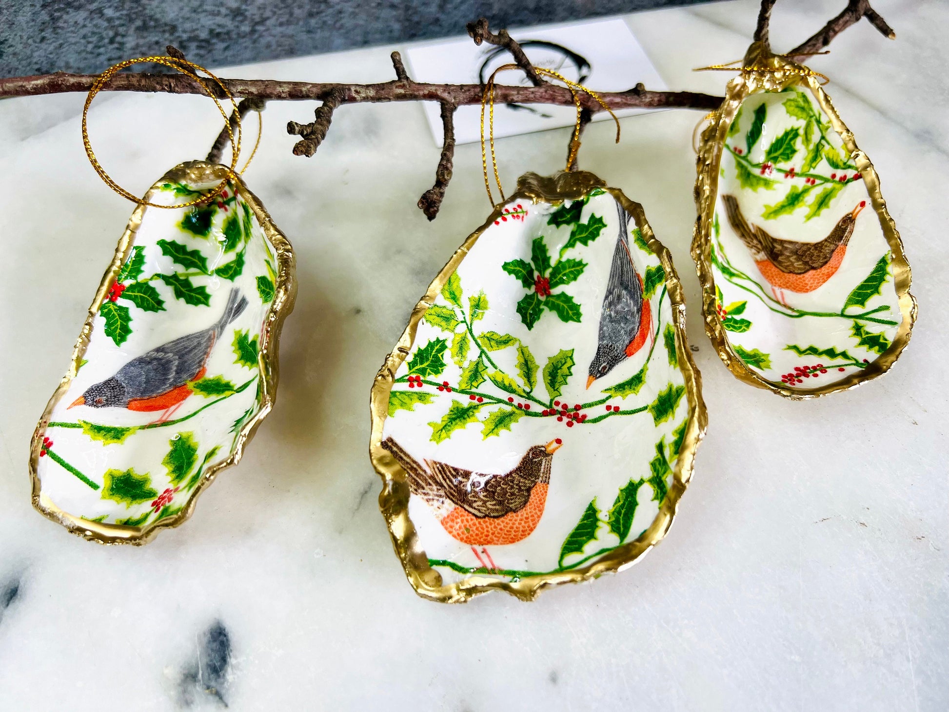 Handmade Dorset oyster shell Christmas ornament featuring red and orange winter birds perched amongst festive green foliage and berries. Coastal oyster shell trinket dish with hand applied gold leaf edging and glossy resin finish, styled as luxury Christmas décor. Sustainable handcrafted robin-inspired shell decoration available as hanging ornament or jewellery dish, inspired by British winter wildlife and the Dorset coastline.