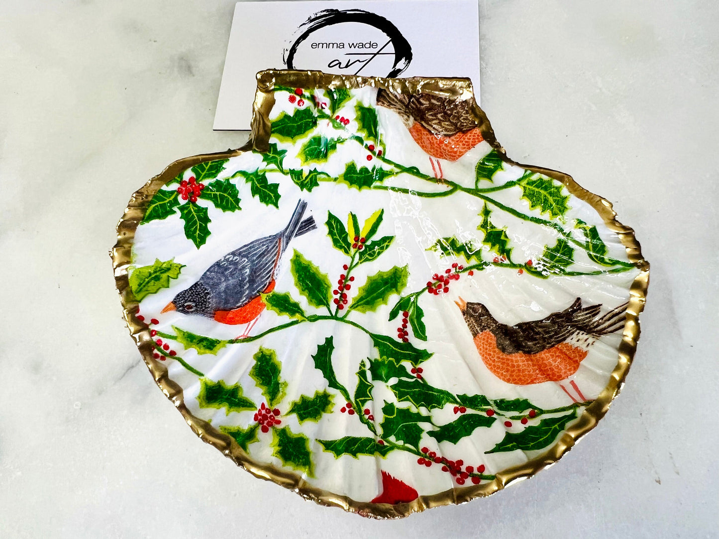 Handmade Dorset scallop shell trinket dish decorated with winter robin and woodland bird illustrations surrounded by festive holly leaves and berries. Natural shell hand finished with liquid gold leaf edging and sealed with glossy resin. Bird décor inspired by coastal cottage interiors and nature styling, suitable as a jewellery dish, ring tray, festive ornament, robin lover gift, winter home décor or Christmas stocking filler.