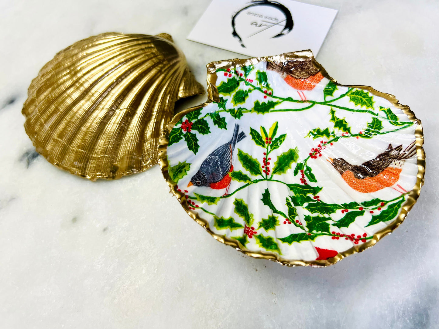 Handmade Dorset scallop shell trinket dish decorated with winter robin and woodland bird illustrations surrounded by festive holly leaves and berries. Natural shell hand finished with liquid gold leaf edging and sealed with glossy resin. Bird décor inspired by coastal cottage interiors and nature styling, suitable as a jewellery dish, ring tray, festive ornament, robin lover gift, winter home décor or Christmas stocking filler.