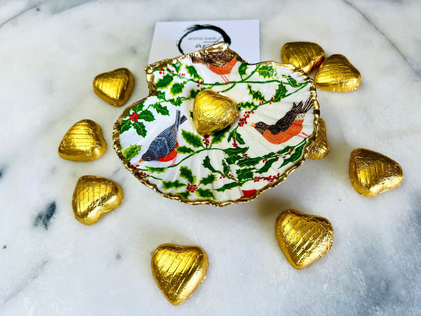Handmade Dorset scallop shell trinket dish decorated with winter robin and woodland bird illustrations surrounded by festive holly leaves and berries. Natural shell hand finished with liquid gold leaf edging and sealed with glossy resin. Bird décor inspired by coastal cottage interiors and nature styling, suitable as a jewellery dish, ring tray, festive ornament, robin lover gift, winter home décor or Christmas stocking filler.