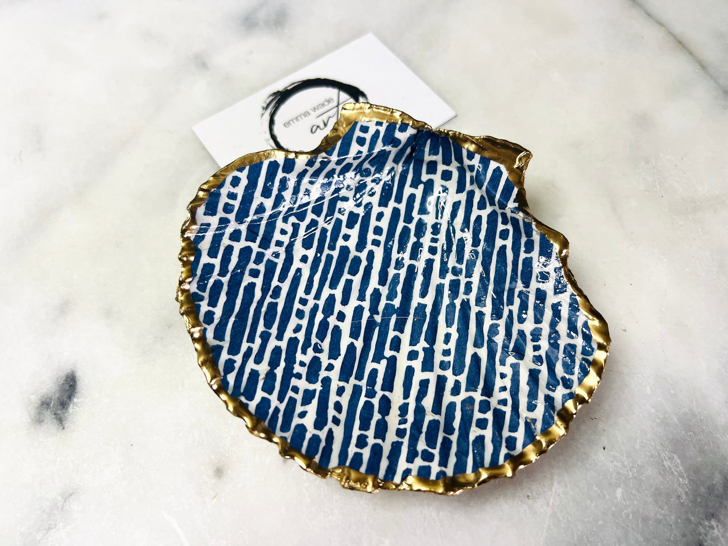 A handmade blue and white scallop shell trinket dish decorated with fine linear detailing and finished with gold leaf around the edge, presented in a sustainable gift box with white tissue paper and shredded packing. The dish is made from a real Dorset scallop shell and sealed with a clear glossy resin, and is suitable for use as a ring dish, jewellery holder, or small decorative tray in a bedroom or bathroom.