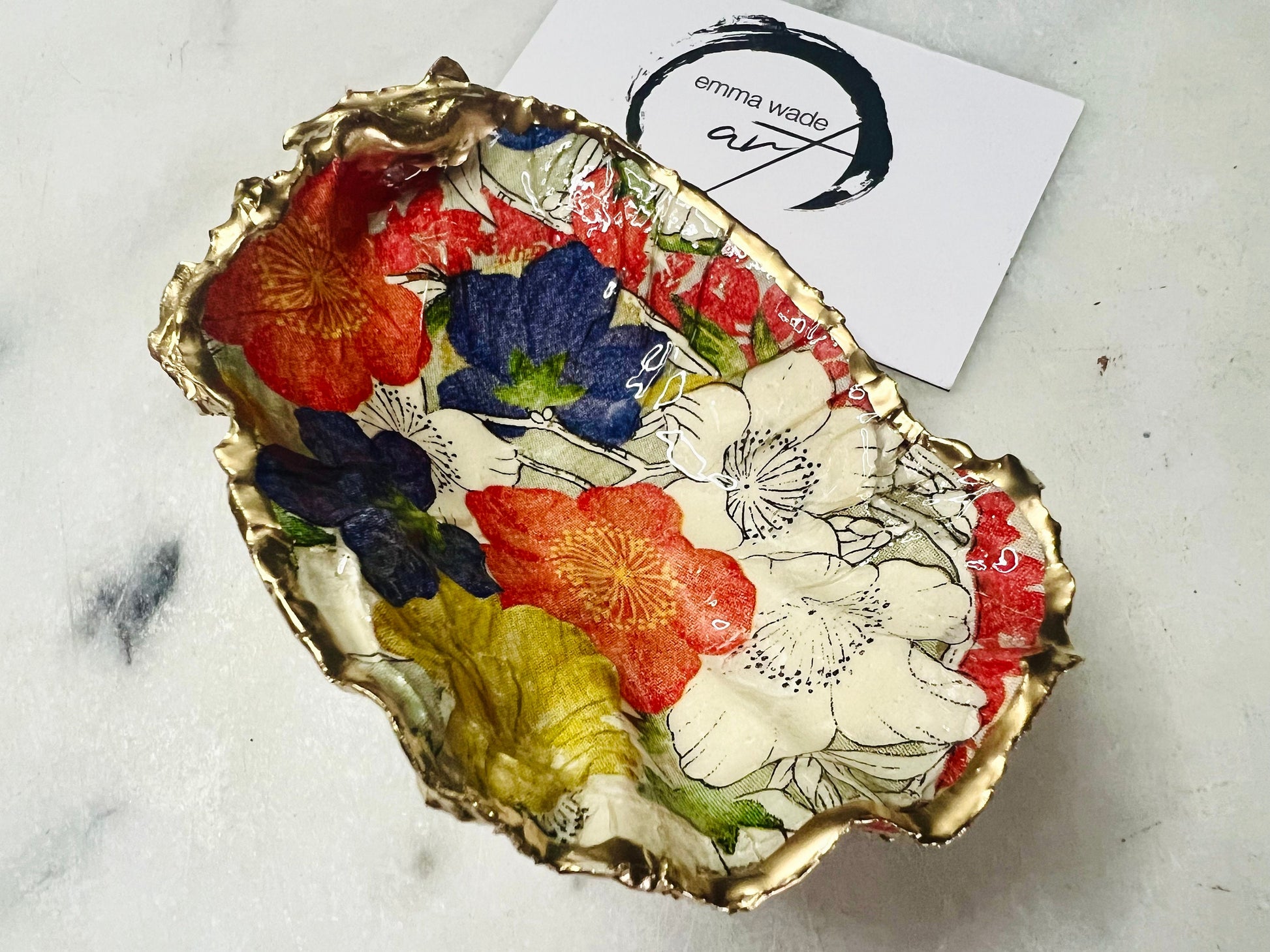 Handmade oyster shell trinket dish featuring a colourful Japanese kimono-style floral design applied with decoupage, finished with hand-gilded gold leaf edges and sealed in glossy clear resin. Natural Dorset oyster shell used as a jewellery dish or ring holder, photographed on a light surface, showcasing the vibrant flowers and luxury gold detailing. Ideal as coastal home decor or a unique handmade gift.