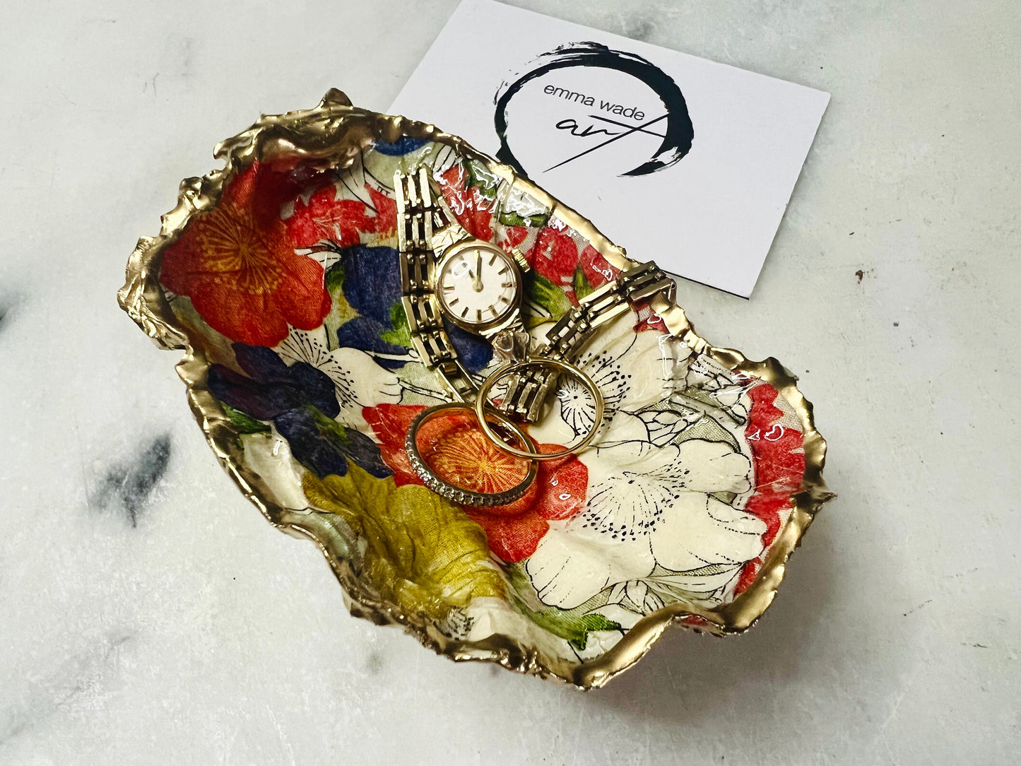 Handmade oyster shell trinket dish featuring a colourful Japanese kimono-style floral design applied with decoupage, finished with hand-gilded gold leaf edges and sealed in glossy clear resin. Natural Dorset oyster shell used as a jewellery dish or ring holder, photographed on a light surface, showcasing the vibrant flowers and luxury gold detailing. Ideal as coastal home decor or a unique handmade gift.