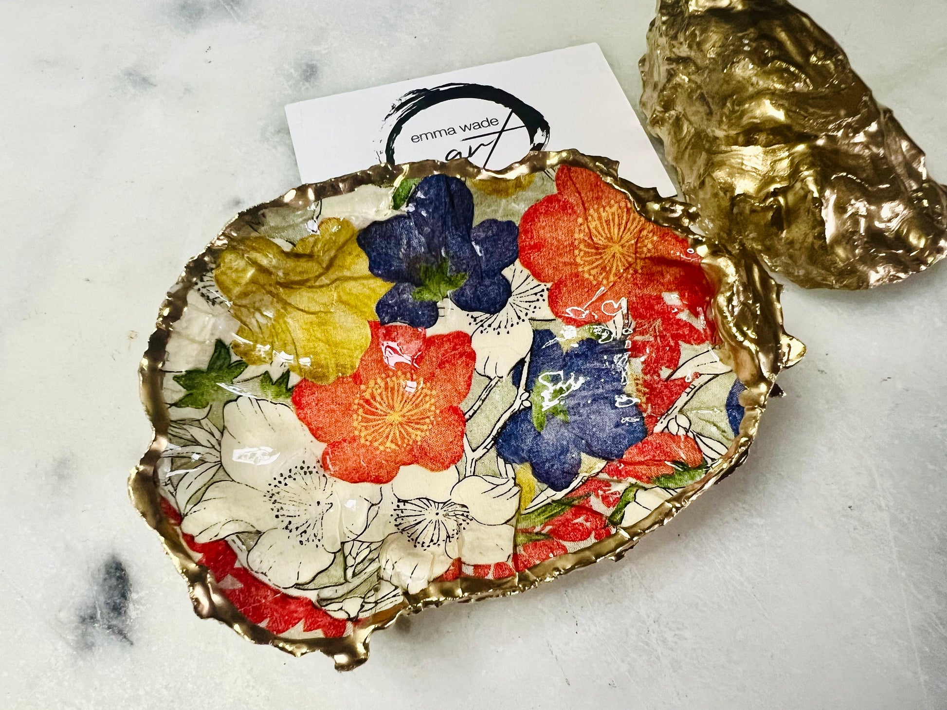 Handmade oyster shell trinket dish featuring a colourful Japanese kimono-style floral design applied with decoupage, finished with hand-gilded gold leaf edges and sealed in glossy clear resin. Natural Dorset oyster shell used as a jewellery dish or ring holder, photographed on a light surface, showcasing the vibrant flowers and luxury gold detailing. Ideal as coastal home decor or a unique handmade gift.