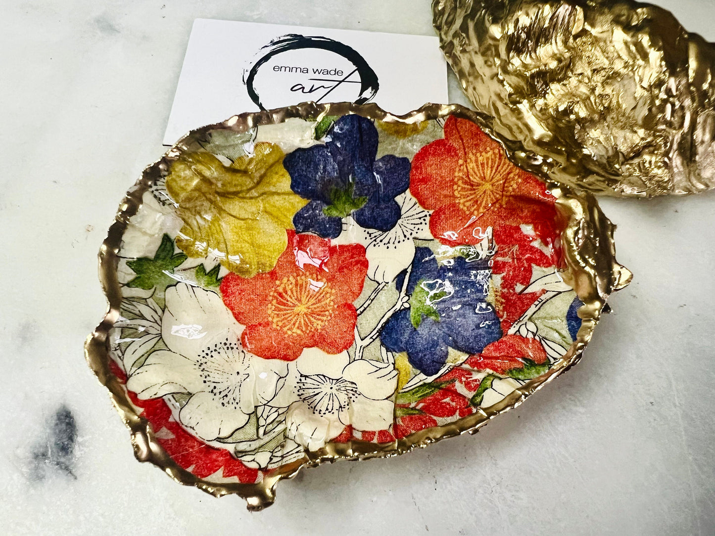 Handmade oyster shell trinket dish featuring a colourful Japanese kimono-style floral design applied with decoupage, finished with hand-gilded gold leaf edges and sealed in glossy clear resin. Natural Dorset oyster shell used as a jewellery dish or ring holder, photographed on a light surface, showcasing the vibrant flowers and luxury gold detailing. Ideal as coastal home decor or a unique handmade gift.