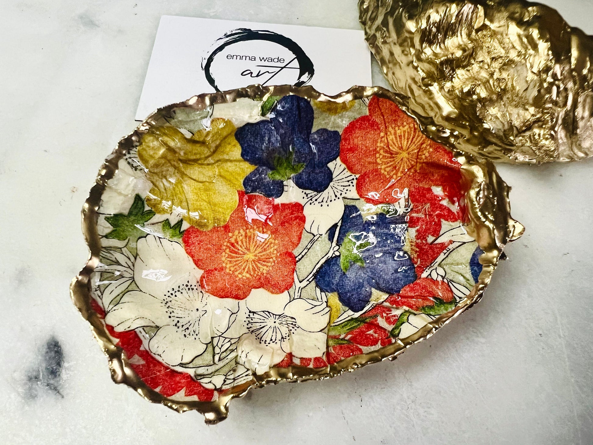 Handmade oyster shell trinket dish featuring a colourful Japanese kimono-style floral design applied with decoupage, finished with hand-gilded gold leaf edges and sealed in glossy clear resin. Natural Dorset oyster shell used as a jewellery dish or ring holder, photographed on a light surface, showcasing the vibrant flowers and luxury gold detailing. Ideal as coastal home decor or a unique handmade gift.