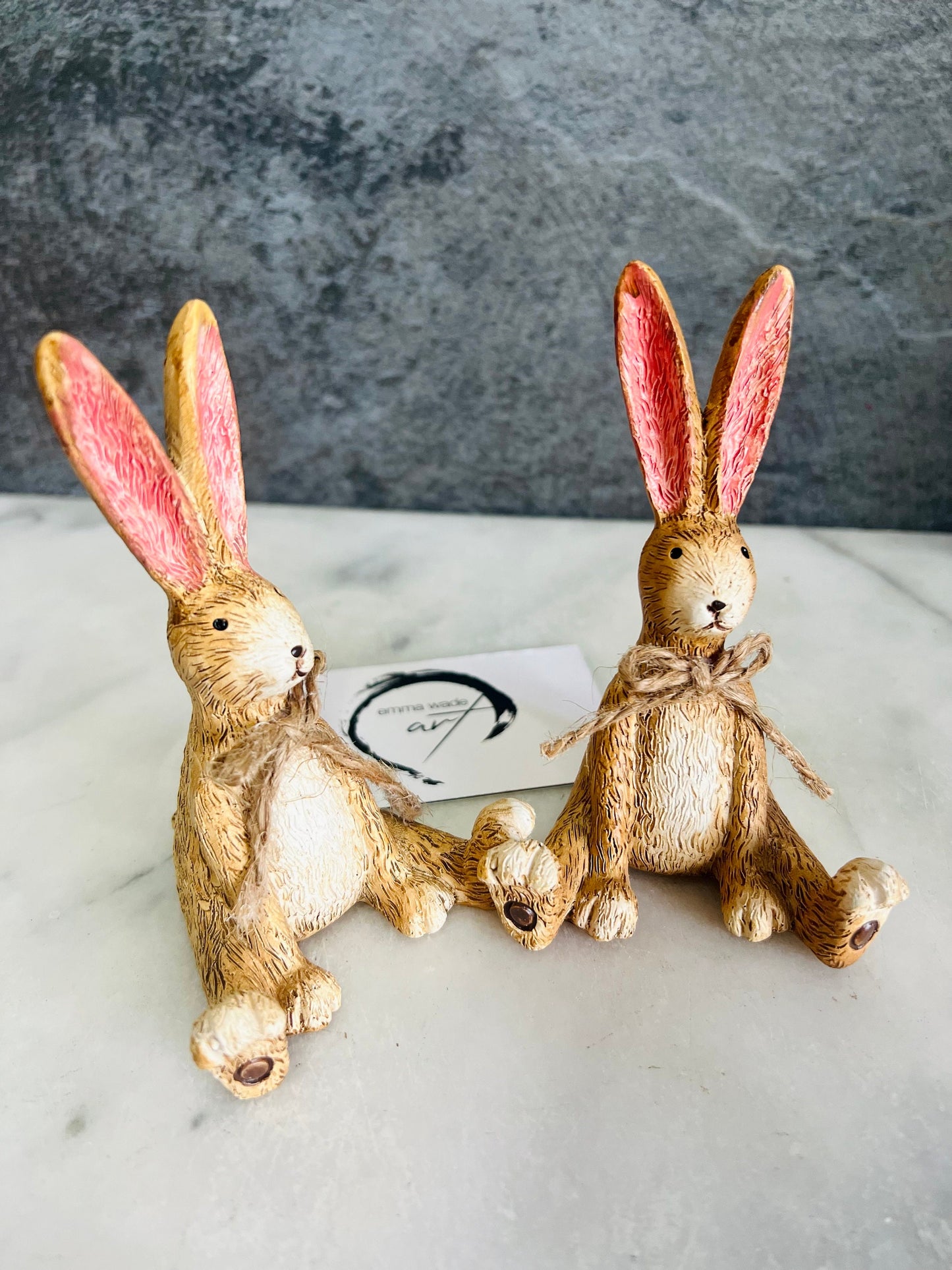Hand finished brown resin rabbit ornament with rustic jute bow and pink inner ears, seated bunny shelf sitter with detailed textured fur finish. Decorative rabbit figurine styled on a wooden stand against a neutral background. A charming rustic home decor piece ideal for mantel display, nursery styling, cottage interiors or country farmhouse decor. Thoughtful gift for her, new home gift, Mother’s Day present or year round bunny decoration.
