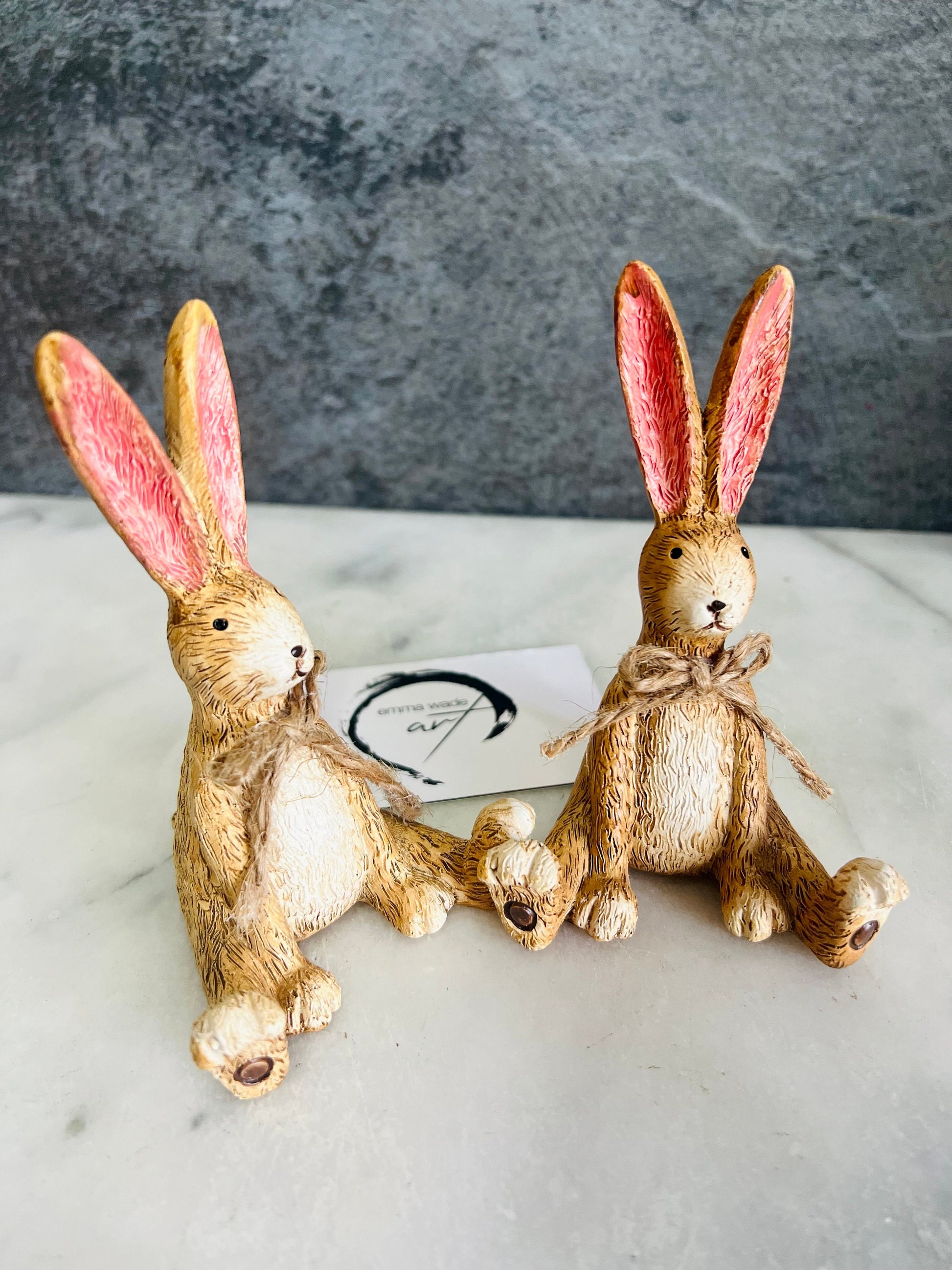 Hand finished brown resin rabbit ornament with rustic jute bow and pink inner ears, seated bunny shelf sitter with detailed textured fur finish. Decorative rabbit figurine styled on a wooden stand against a neutral background. A charming rustic home decor piece ideal for mantel display, nursery styling, cottage interiors or country farmhouse decor. Thoughtful gift for her, new home gift, Mother’s Day present or year round bunny decoration.