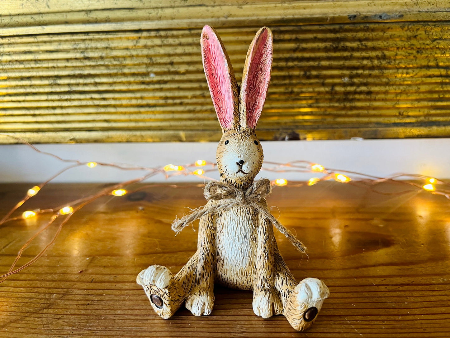 Hand finished brown resin rabbit ornament with rustic jute bow and pink inner ears, seated bunny shelf sitter with detailed textured fur finish. Decorative rabbit figurine styled on a wooden stand against a neutral background. A charming rustic home decor piece ideal for mantel display, nursery styling, cottage interiors or country farmhouse decor. Thoughtful gift for her, new home gift, Mother’s Day present or year round bunny decoration.