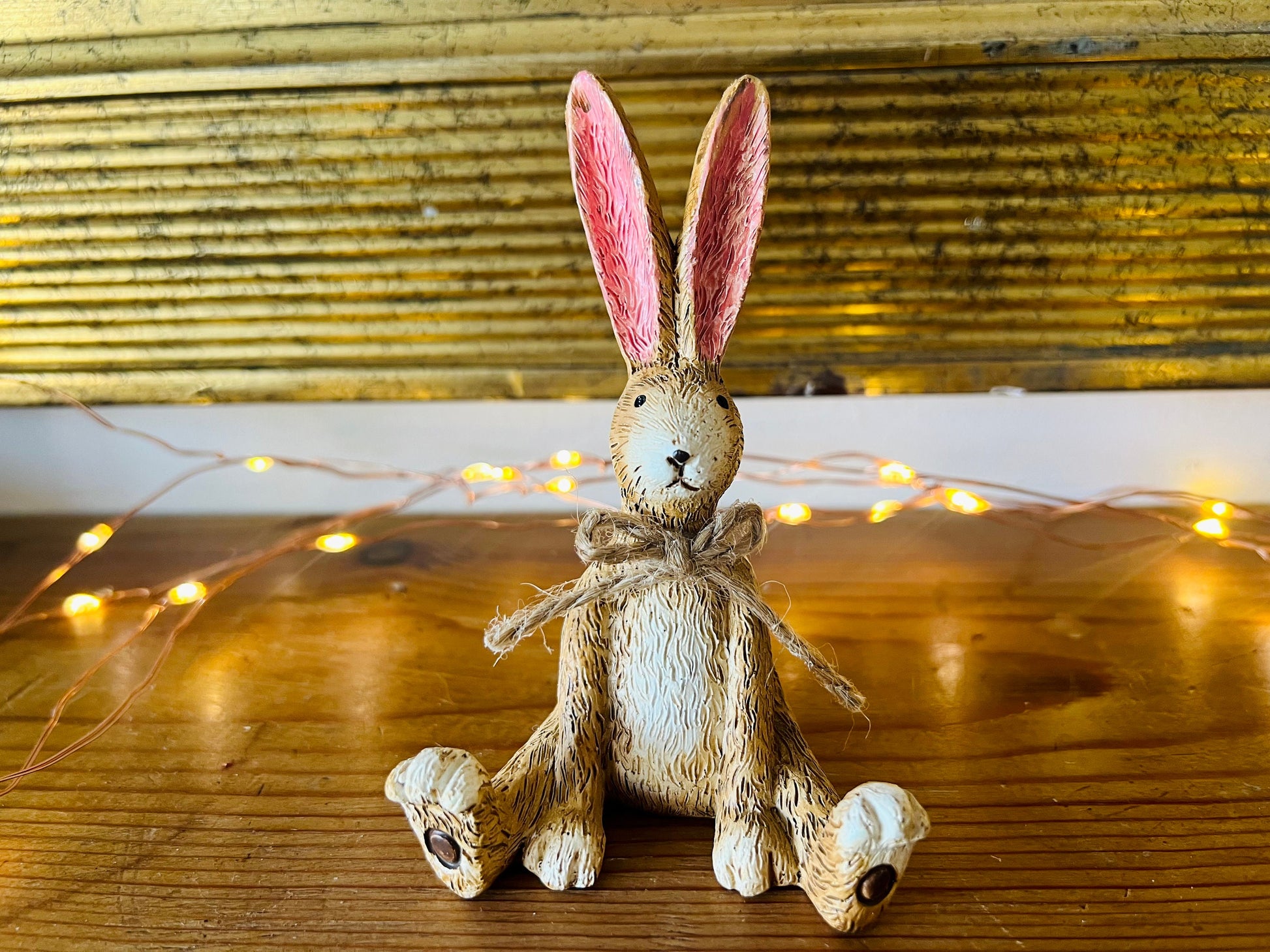 Hand finished brown resin rabbit ornament with rustic jute bow and pink inner ears, seated bunny shelf sitter with detailed textured fur finish. Decorative rabbit figurine styled on a wooden stand against a neutral background. A charming rustic home decor piece ideal for mantel display, nursery styling, cottage interiors or country farmhouse decor. Thoughtful gift for her, new home gift, Mother’s Day present or year round bunny decoration.