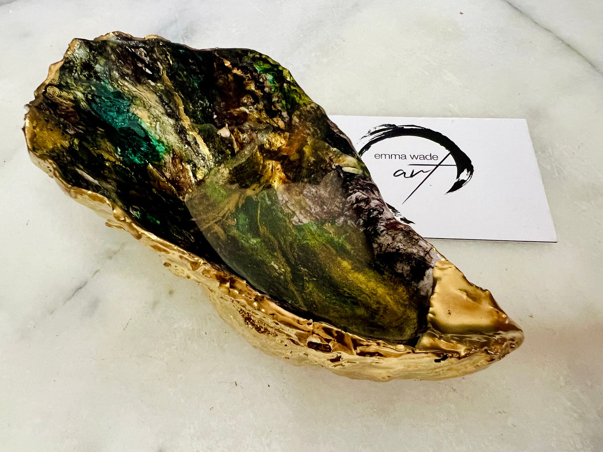 Extra large oyster shell dish with green and gold lagoon design, created using poured acrylic inks and layered resins for a deep ocean effect. Finished with shimmering gold edges, this handmade shell is a unique one-of-a-kind piece of coastal art. Can be used as a jewellery dish, ring holder, trinket tray, or decorative ornament. Also suitable as a serving dish for canapés or chocolates. Inspired by sea caves and lagoons, each shell is natural, rare, and individually crafted.
