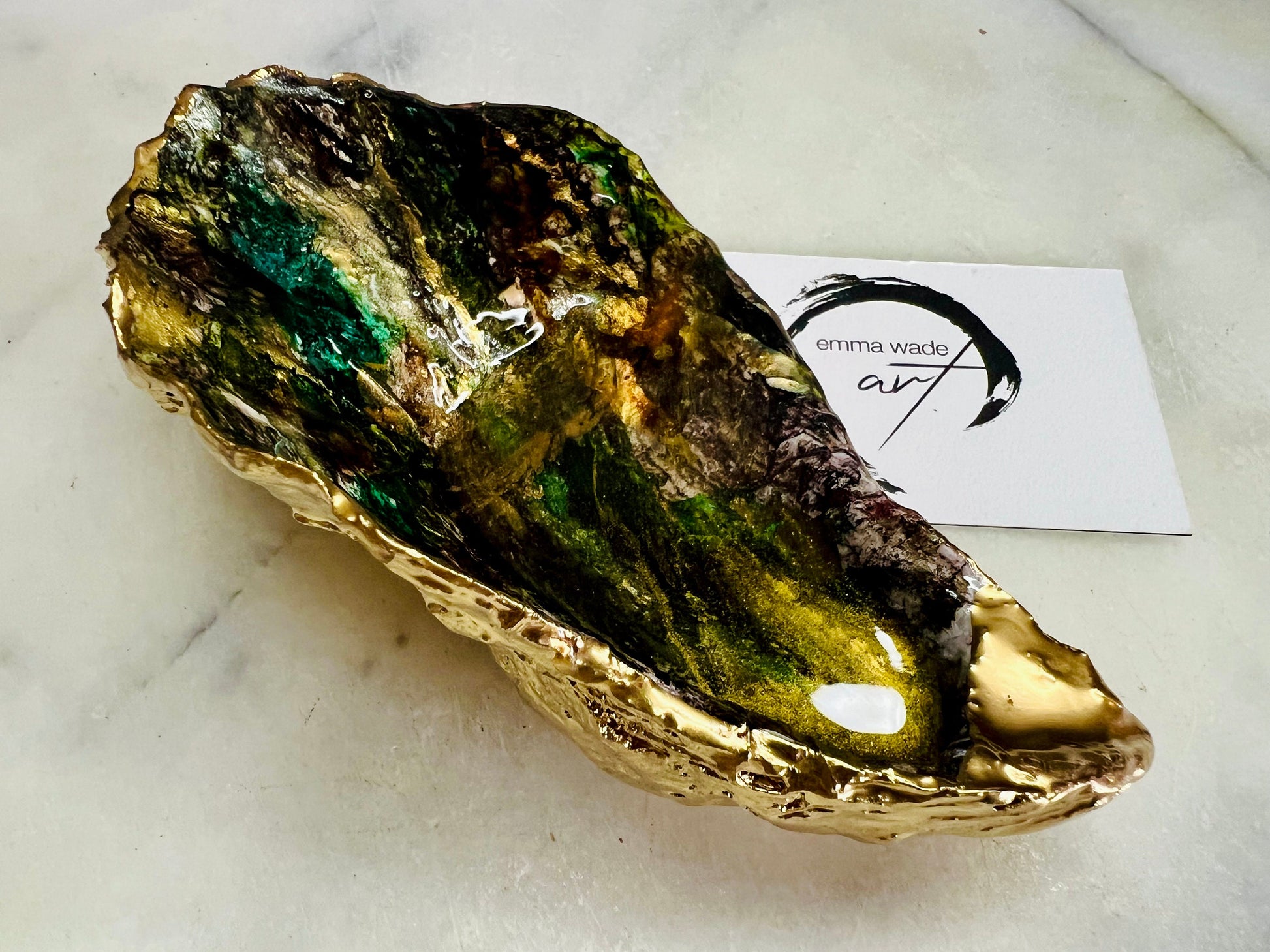 Extra large oyster shell dish with green and gold lagoon design, created using poured acrylic inks and layered resins for a deep ocean effect. Finished with shimmering gold edges, this handmade shell is a unique one-of-a-kind piece of coastal art. Can be used as a jewellery dish, ring holder, trinket tray, or decorative ornament. Also suitable as a serving dish for canapés or chocolates. Inspired by sea caves and lagoons, each shell is natural, rare, and individually crafted.