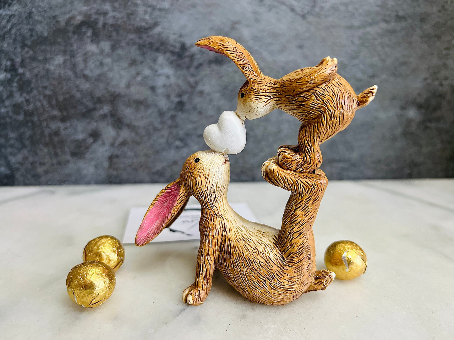 Hand finished brown polyresin bunny and baby rabbit ornament, showing a larger rabbit balancing a baby bunny above with a white heart between them. Rustic textured rabbit figure with pink inner ears, suitable for Christmas shelf decor, mantel styling, rabbit lover gifts and year-round home decoration.