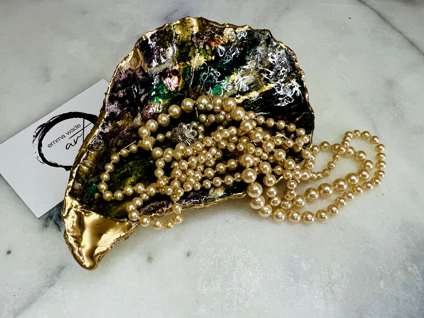 Extra large oyster shell dish with green and gold lagoon design, created using poured acrylic inks and layered resins for a deep ocean effect. Finished with shimmering gold edges, this handmade shell is a unique one-of-a-kind piece of coastal art. Can be used as a jewellery dish, ring holder, trinket tray, or decorative ornament. Also suitable as a serving dish for canapés or chocolates. Inspired by sea caves and lagoons, each shell is natural, rare, and individually crafted.