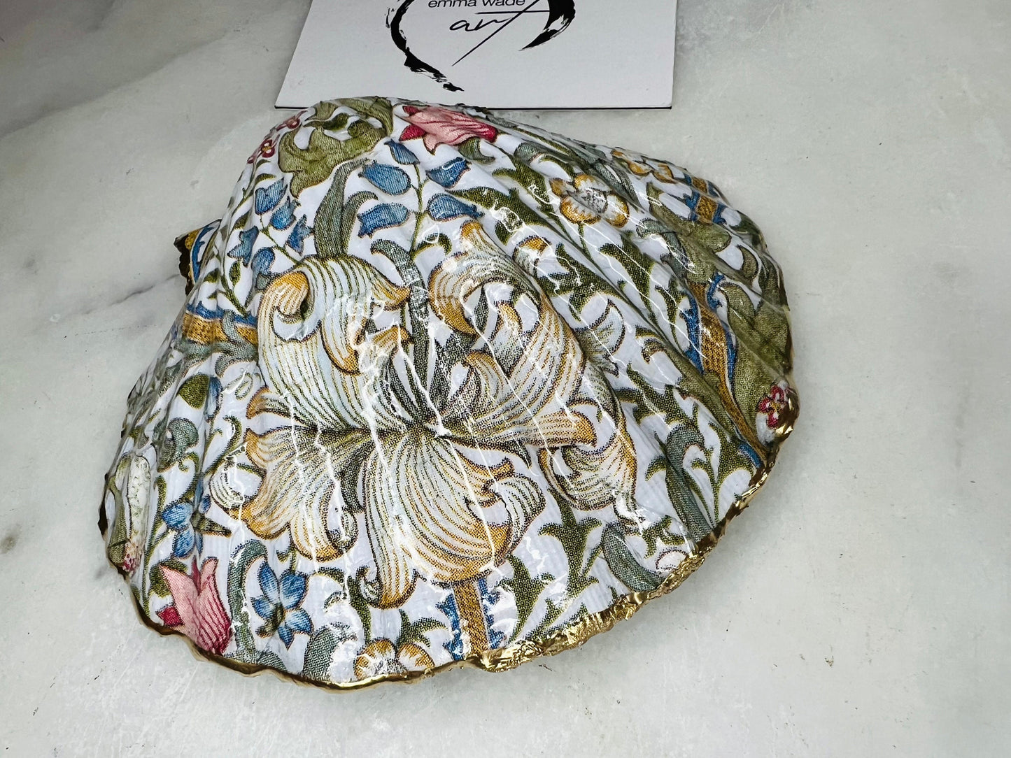 Handcrafted Dorset scallop shell trinket dish featuring William Morris Golden Lily floral design. Natural shell edged with hand applied gold leaf and sealed with glossy resin. Heritage floral jewellery holder displayed with rings on marble surface. Elegant coastal home decor gift.