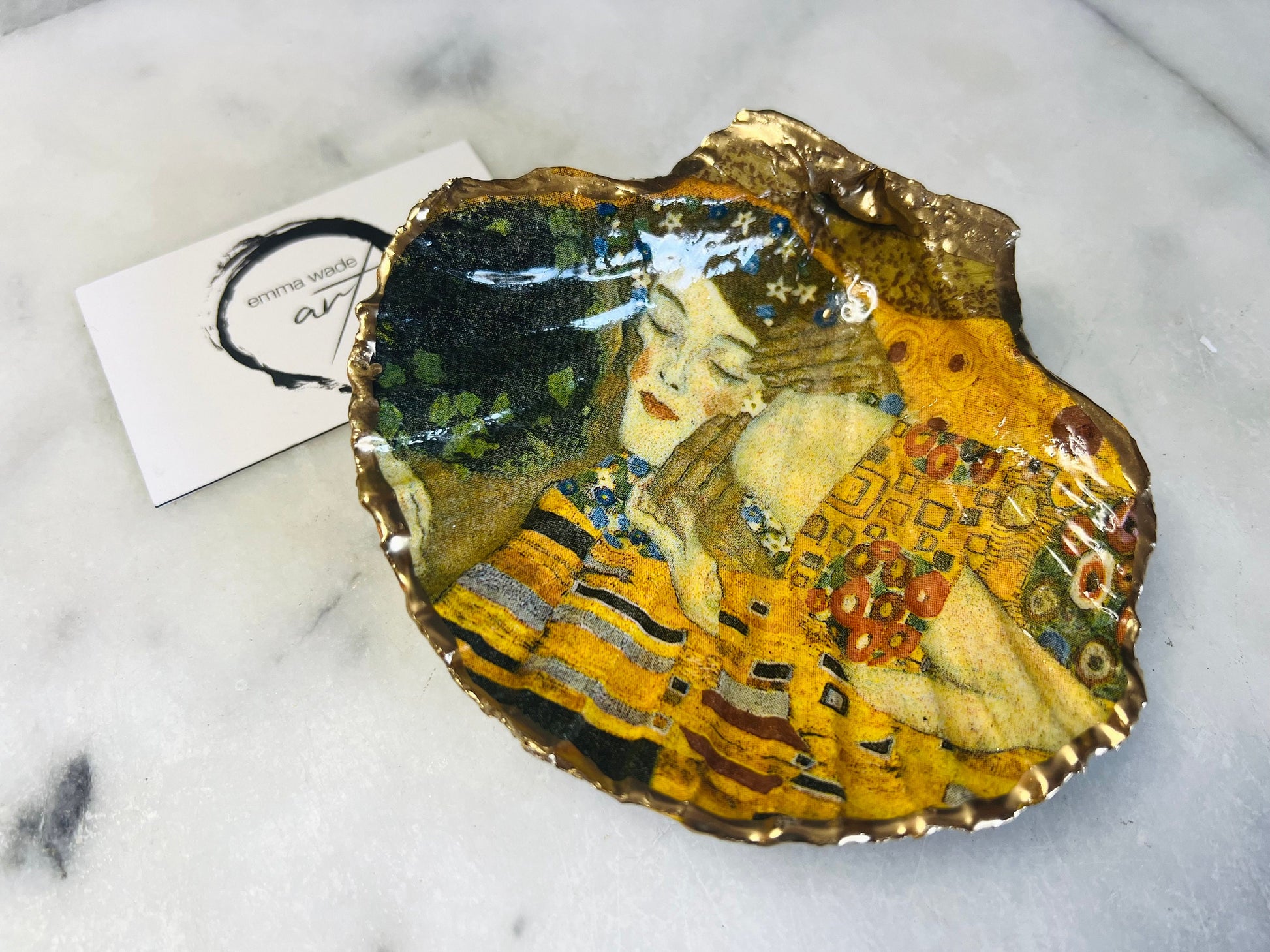 Handmade scallop shell trinket dish inspired by Gustav Klimt’s “The Kiss”, finished with gold leaf and sealed with glossy resin. Coastal Dorset shell jewellery dish perfect for rings, earrings and small treasures. Unique decorative shell bowl with golden artwork design, photographed on a light background showing detailed gilded surface and natural shell shape.