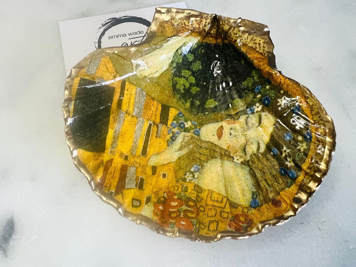 Handmade scallop shell trinket dish inspired by Gustav Klimt’s “The Kiss”, finished with gold leaf and sealed with glossy resin. Coastal Dorset shell jewellery dish perfect for rings, earrings and small treasures. Unique decorative shell bowl with golden artwork design, photographed on a light background showing detailed gilded surface and natural shell shape.