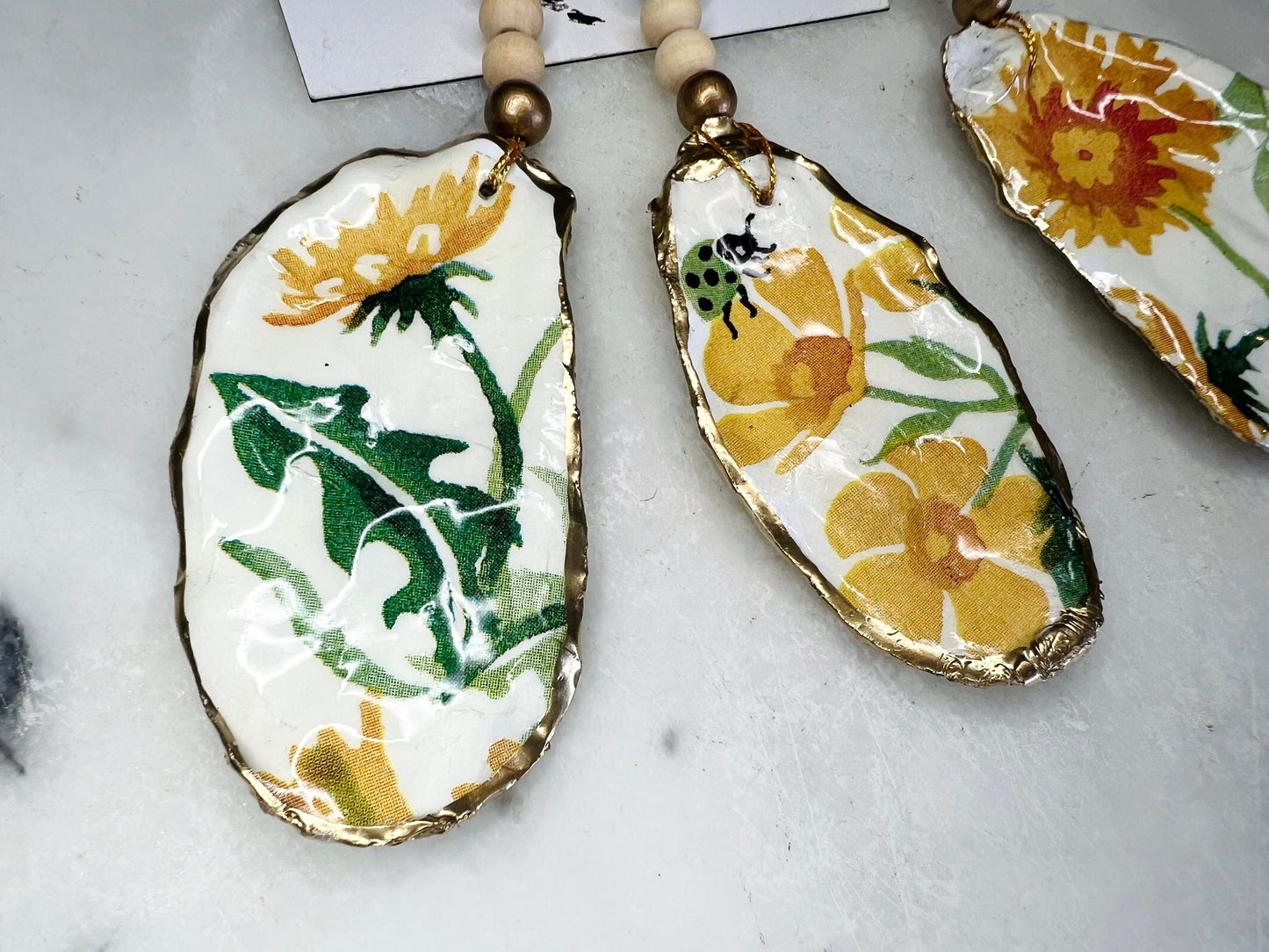Handmade Dorset oyster shell hanging ornament featuring genuine Emma Bridgewater yellow spring floral napkin design, finished with gold leaf edging and glossy resin. Natural pearlescent reverse with gold detailing and gold hanging cord with decorative beads. Coastal seasonal home decor.