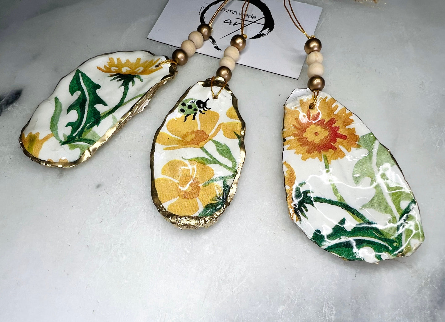 Handmade Dorset oyster shell hanging ornament featuring genuine Emma Bridgewater yellow spring floral napkin design, finished with gold leaf edging and glossy resin. Natural pearlescent reverse with gold detailing and gold hanging cord with decorative beads. Coastal seasonal home decor.