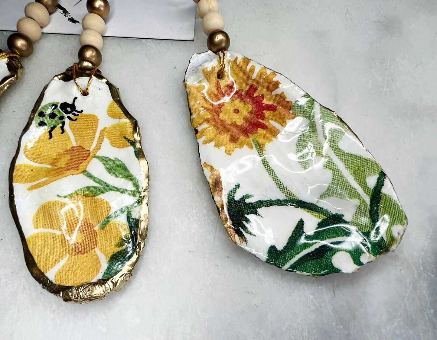 Handmade Dorset oyster shell hanging ornament featuring genuine Emma Bridgewater yellow spring floral napkin design, finished with gold leaf edging and glossy resin. Natural pearlescent reverse with gold detailing and gold hanging cord with decorative beads. Coastal seasonal home decor.