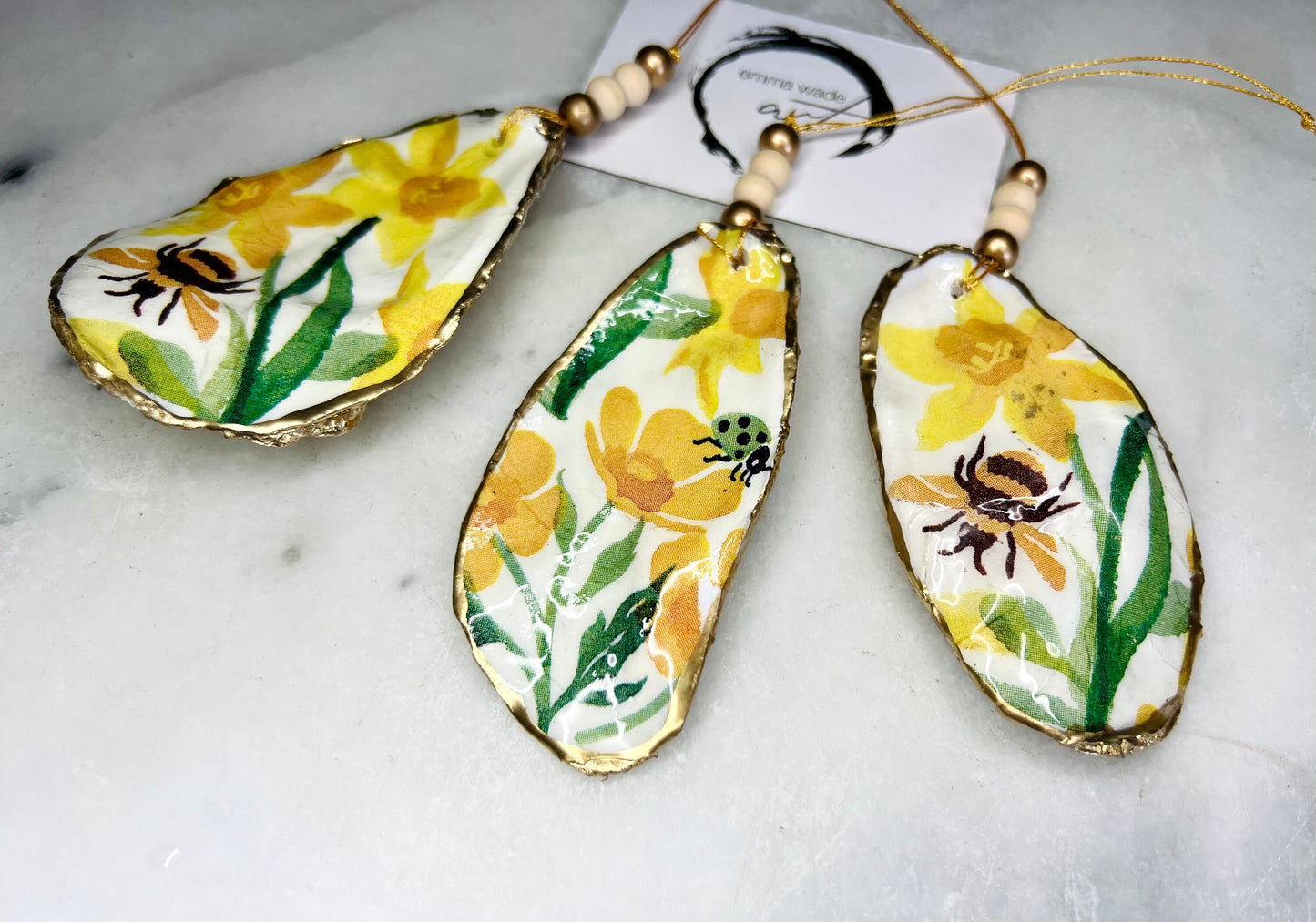 Handcrafted Dorset oyster shell hanging ornament featuring genuine Emma Bridgewater yellow spring floral napkin design with bright yellow flowers, green foliage, bumblebees and ladybirds. Finished with liquid gold leaf edging and sealed in glossy resin. Natural pearlescent reverse with gold detailing and gold hanging cord with decorative beads. Coastal seasonal home decor.