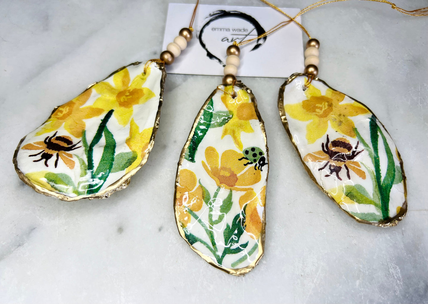 Handmade Dorset oyster shell hanging ornament featuring genuine Emma Bridgewater yellow spring floral napkin design, Bumble bee design, ladybird design, finished with gold leaf edging and glossy resin. Natural pearlescent reverse with gold detailing and gold hanging cord with decorative beads. Coastal seasonal home decor.