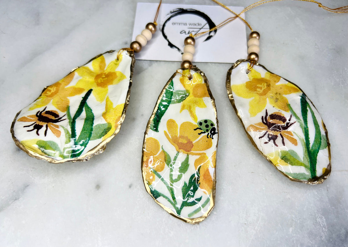 Handmade Dorset oyster shell hanging ornament featuring genuine Emma Bridgewater yellow spring floral napkin design, Bumble bee design, ladybird design, finished with gold leaf edging and glossy resin. Natural pearlescent reverse with gold detailing and gold hanging cord with decorative beads. Coastal seasonal home decor.