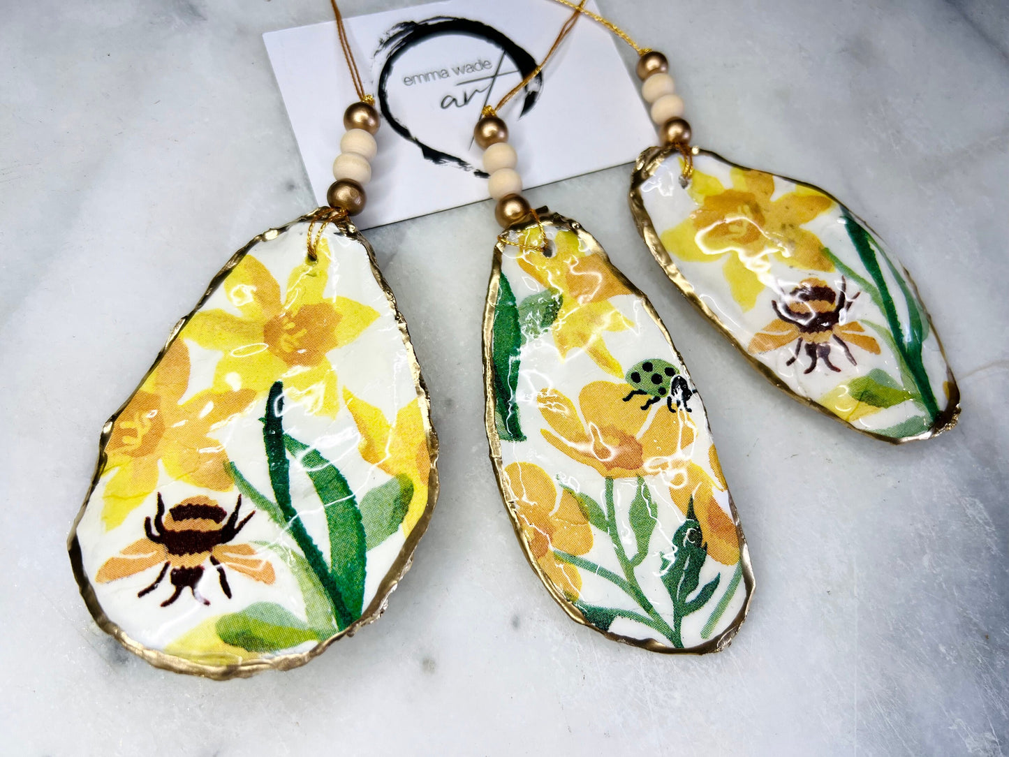 Handcrafted Dorset oyster shell hanging ornament featuring genuine Emma Bridgewater yellow spring floral napkin design with bright yellow flowers, green foliage, bumblebees and ladybirds. Finished with liquid gold leaf edging and sealed in glossy resin. Natural pearlescent reverse with gold detailing and gold hanging cord with decorative beads. Coastal seasonal home decor.