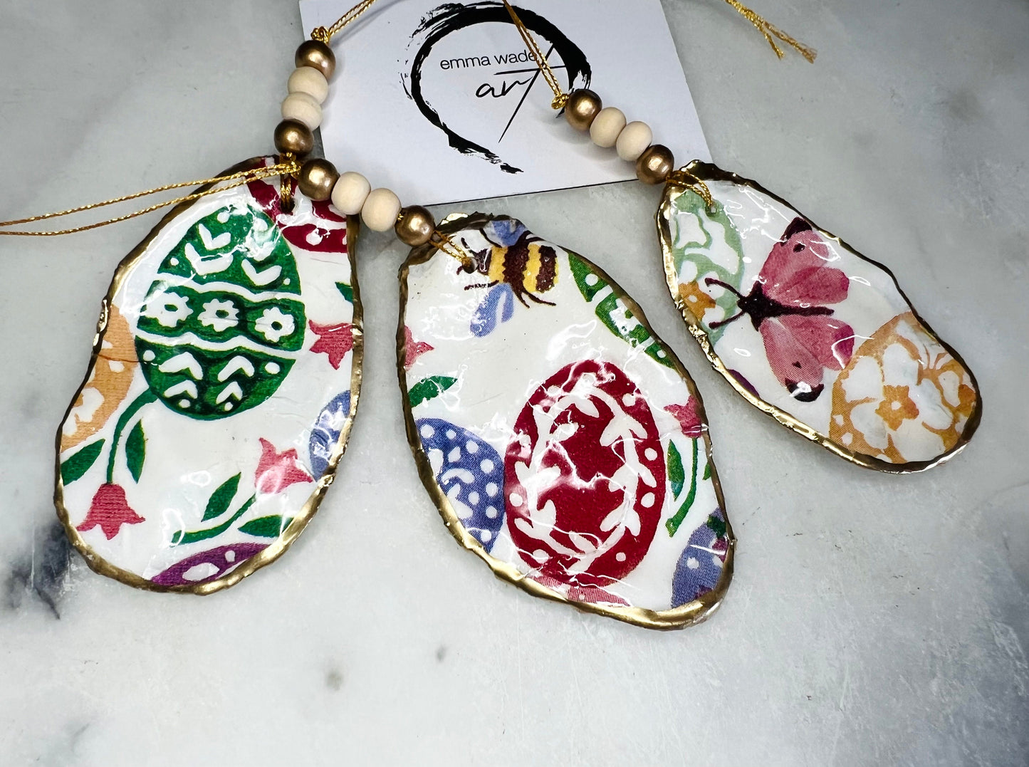Handmade Dorset oyster shell ornaments featuring Emma Bridgewater bee, butterfly, and egg designs. Each shell has a natural pearl finish, gilded with liquid gold leaf on the rim and back, and sealed with glossy resin for shine and durability. Available individually or as a set of 3, with pre-drilled holes, gold hanging string, and decorative beads. Unique festive decorations for Christmas trees, seasonal home décor, or handmade coastal-inspired gifts for weddings, birthdays, or anniversaries.