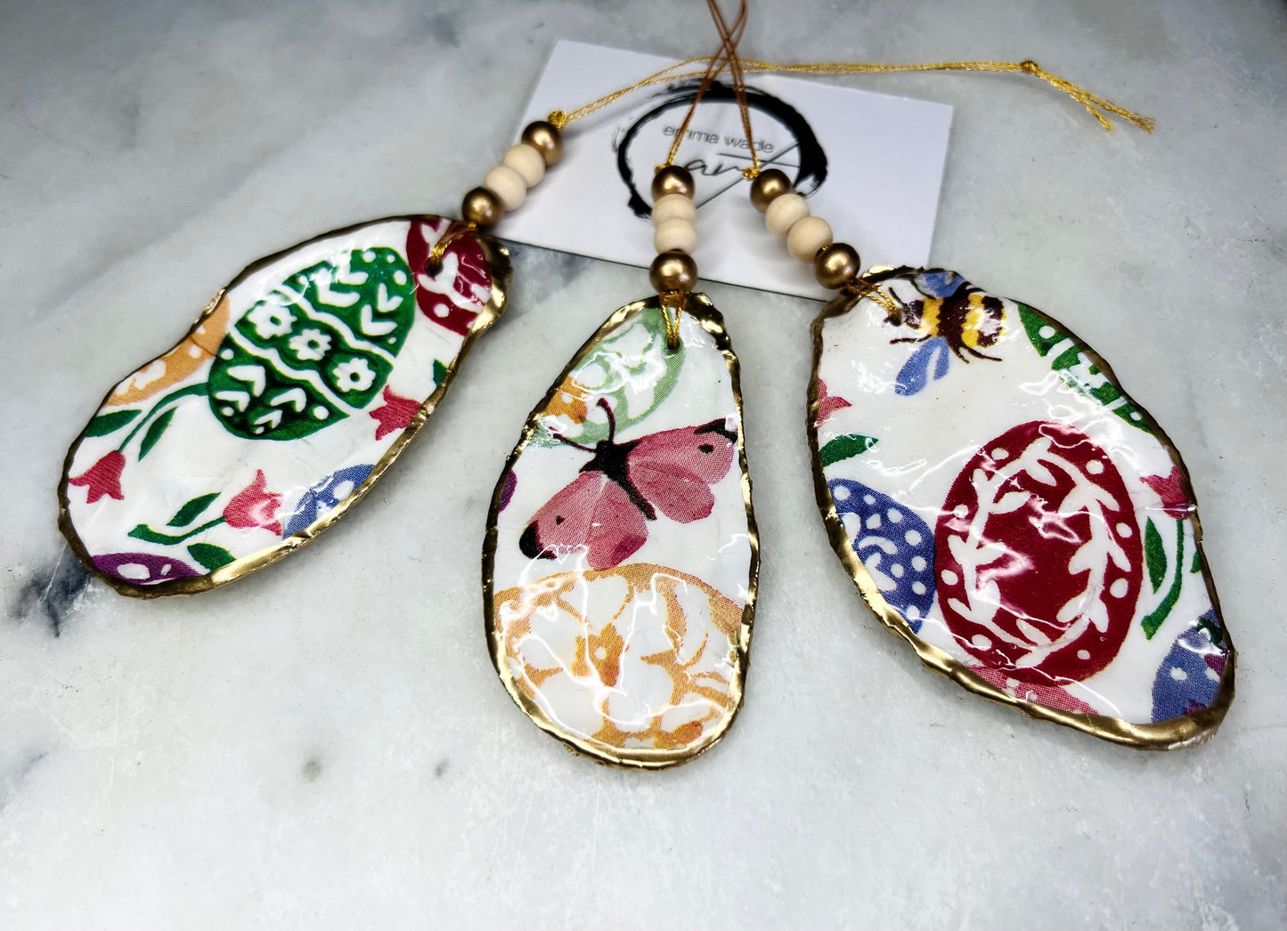 Handmade Dorset oyster shell ornaments featuring Emma Bridgewater bee, butterfly, and egg designs. Each shell has a natural pearl finish, gilded with liquid gold leaf on the rim and back, and sealed with glossy resin for shine and durability. Available individually or as a set of 3, with pre-drilled holes, gold hanging string, and decorative beads. Unique festive decorations for Christmas trees, seasonal home décor, or handmade coastal-inspired gifts for weddings, birthdays, or anniversaries.