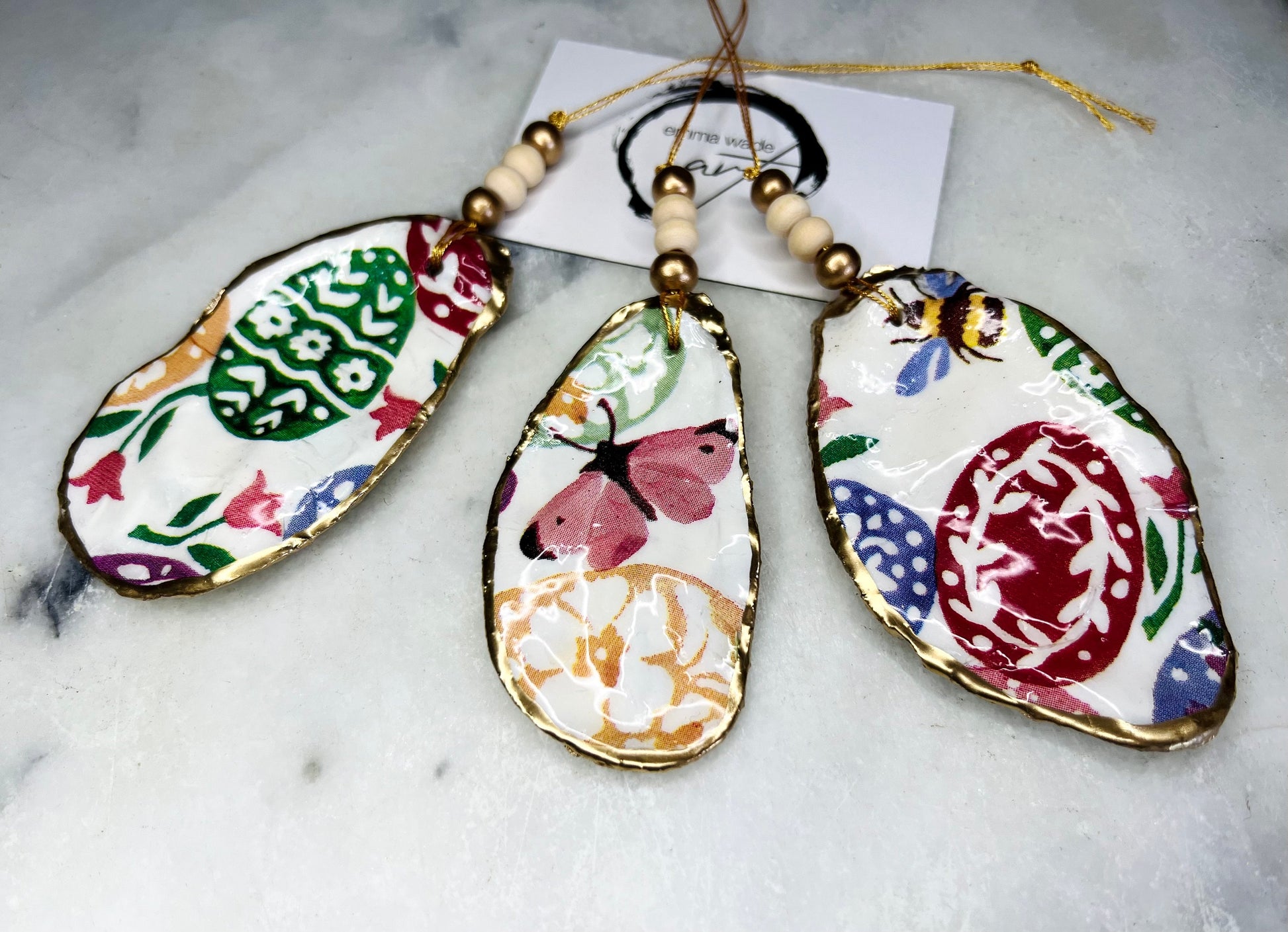 Handmade Dorset oyster shell ornaments featuring Emma Bridgewater bee, butterfly, and egg designs. Each shell has a natural pearl finish, gilded with liquid gold leaf on the rim and back, and sealed with glossy resin for shine and durability. Available individually or as a set of 3, with pre-drilled holes, gold hanging string, and decorative beads. Unique festive decorations for Christmas trees, seasonal home décor, or handmade coastal-inspired gifts for weddings, birthdays, or anniversaries.