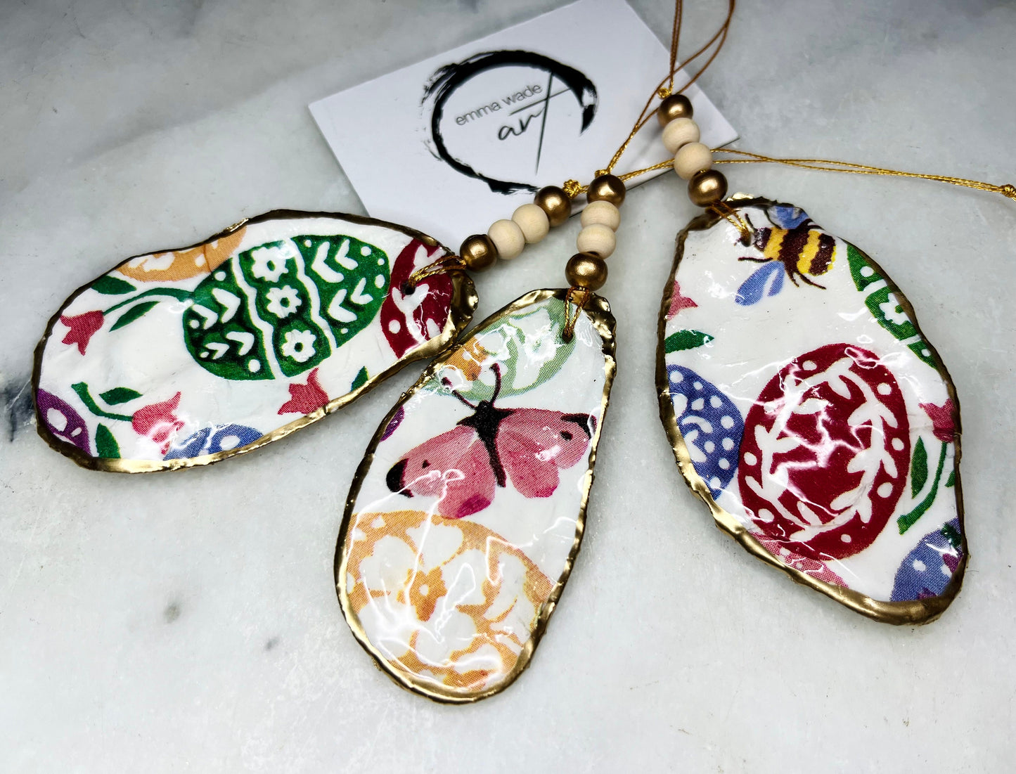 Handmade Dorset oyster shell ornaments featuring Emma Bridgewater bee, butterfly, and egg designs. Each shell has a natural pearl finish, gilded with liquid gold leaf on the rim and back, and sealed with glossy resin for shine and durability. Available individually or as a set of 3, with pre-drilled holes, gold hanging string, and decorative beads. Unique festive decorations for Christmas trees, seasonal home décor, or handmade coastal-inspired gifts for weddings, birthdays, or anniversaries.