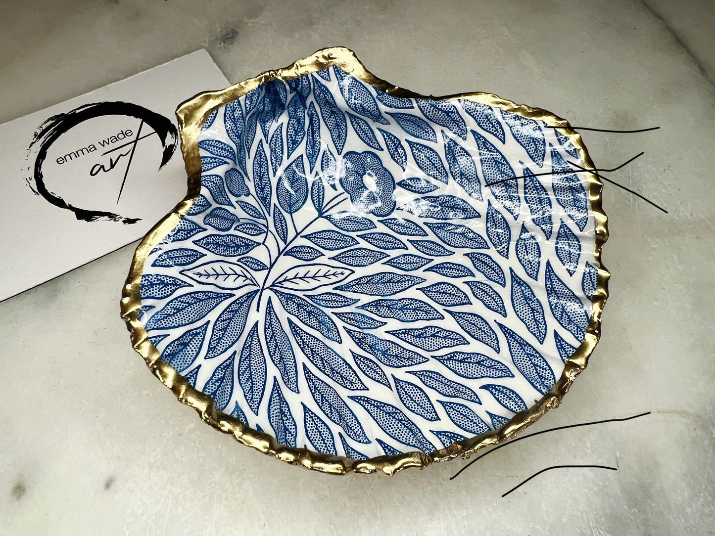 Handmade Dorset scallop shell trinket dishes featuring intricate blue floral and leaf artwork with hand-painted gold leaf edging and glossy crystal clear resin. Coastal botanical jewellery dishes inspired by Nordic Scandinavian decor, styled on a neutral background with elegant blue flower detailing and gilded edges.