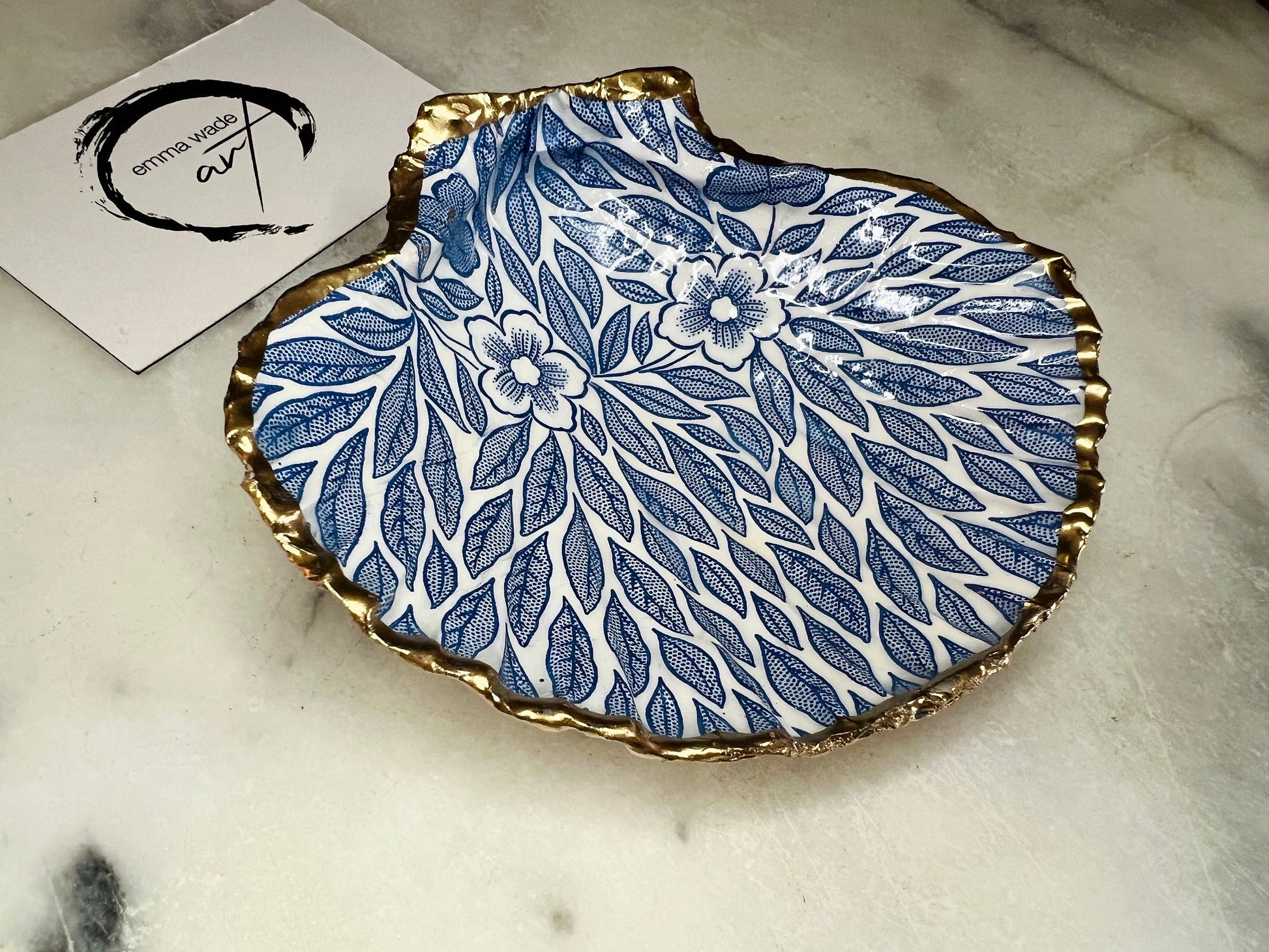 Handmade Dorset scallop shell trinket dishes featuring intricate blue floral and leaf artwork with hand-painted gold leaf edging and glossy crystal clear resin. Coastal botanical jewellery dishes inspired by Nordic Scandinavian decor, styled on a neutral background with elegant blue flower detailing and gilded edges.