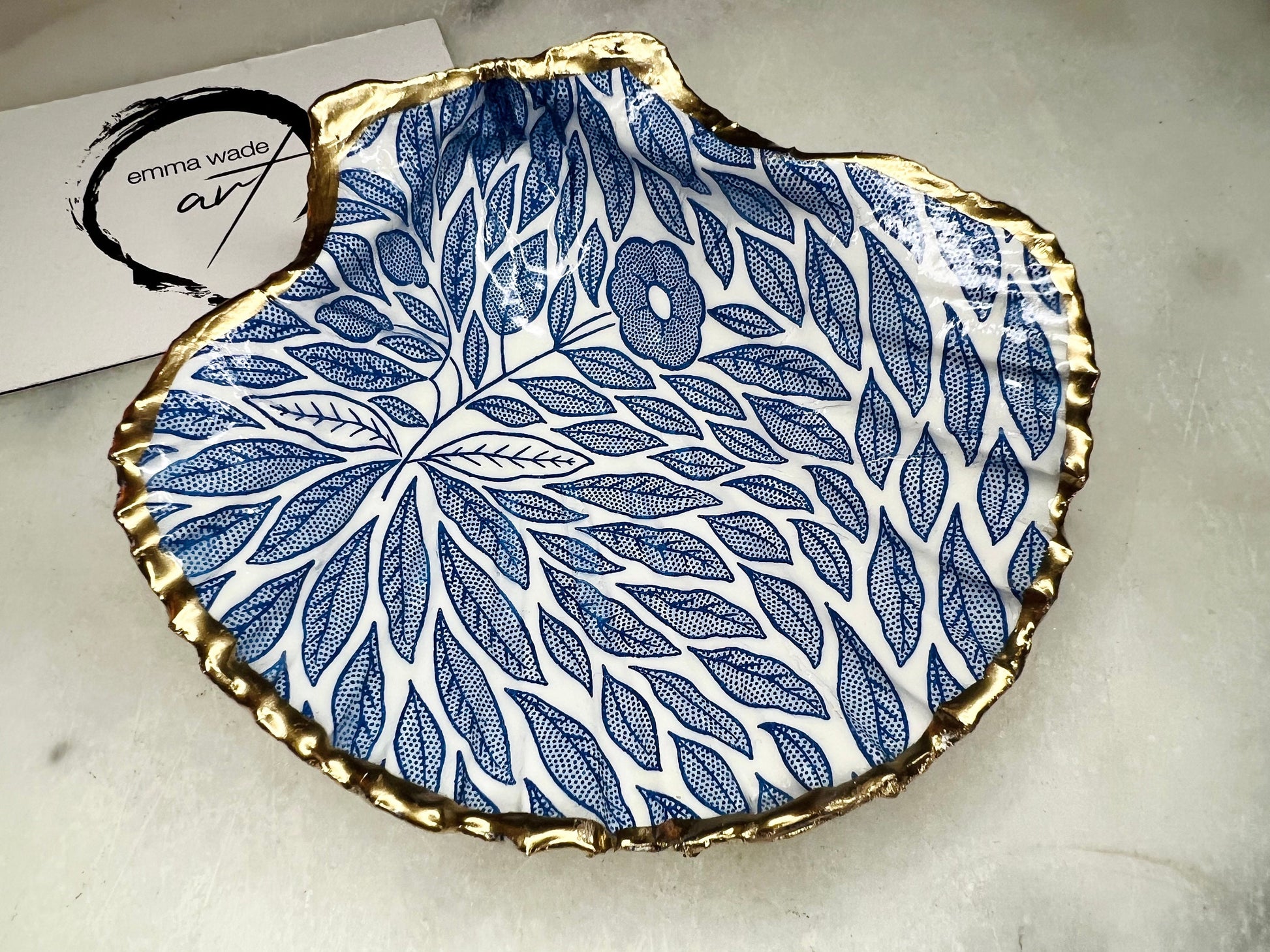 Handmade Dorset scallop shell trinket dishes featuring intricate blue floral and leaf artwork with hand-painted gold leaf edging and glossy crystal clear resin. Coastal botanical jewellery dishes inspired by Nordic Scandinavian decor, styled on a neutral background with elegant blue flower detailing and gilded edges.