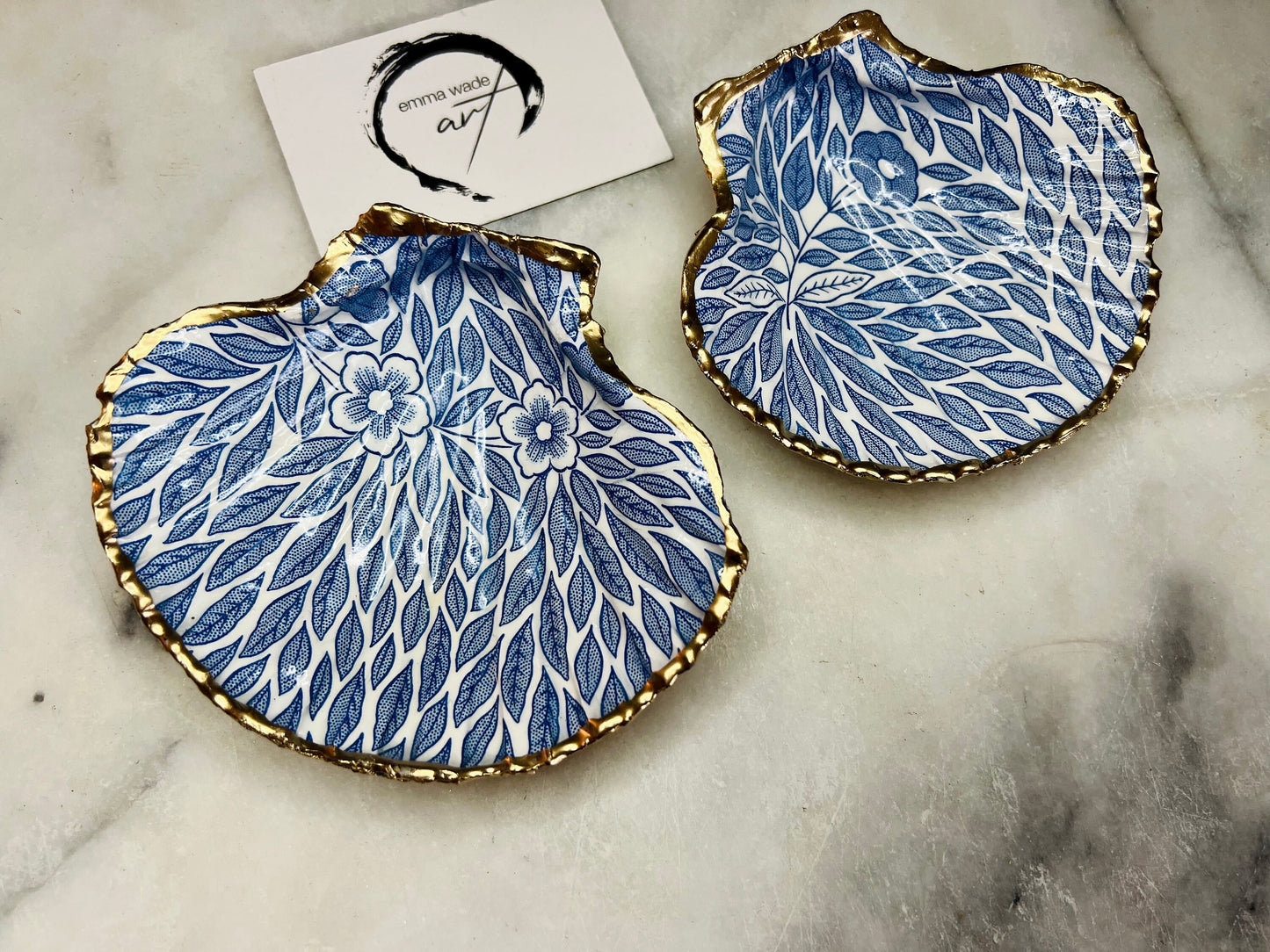 Handmade Dorset scallop shell trinket dishes featuring intricate blue floral and leaf artwork with hand-painted gold leaf edging and glossy crystal clear resin. Coastal botanical jewellery dishes inspired by Nordic Scandinavian decor, styled on a neutral background with elegant blue flower detailing and gilded edges.
