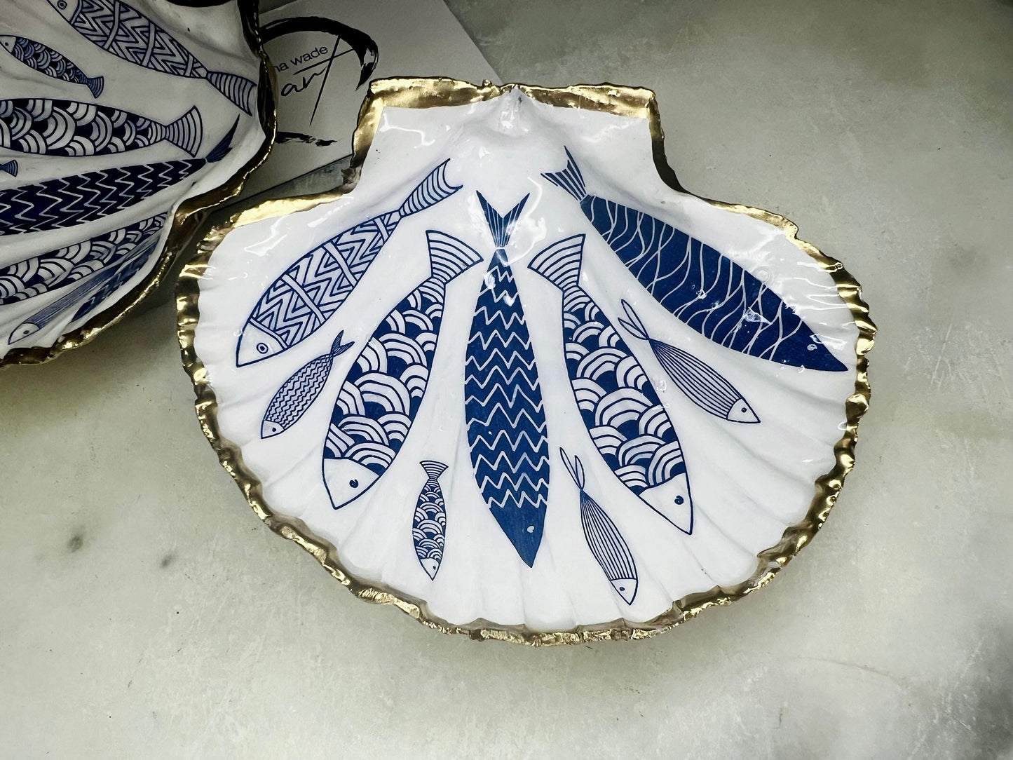 Handmade Dorset scallop shell trinket dishes featuring elegant blue fish illustrations with hand-painted gold leaf edging and glossy crystal clear resin. Coastal nautical jewellery dishes inspired by Scandinavian ocean decor, styled on a neutral background with detailed blue fish artwork and luxurious gilded edges.