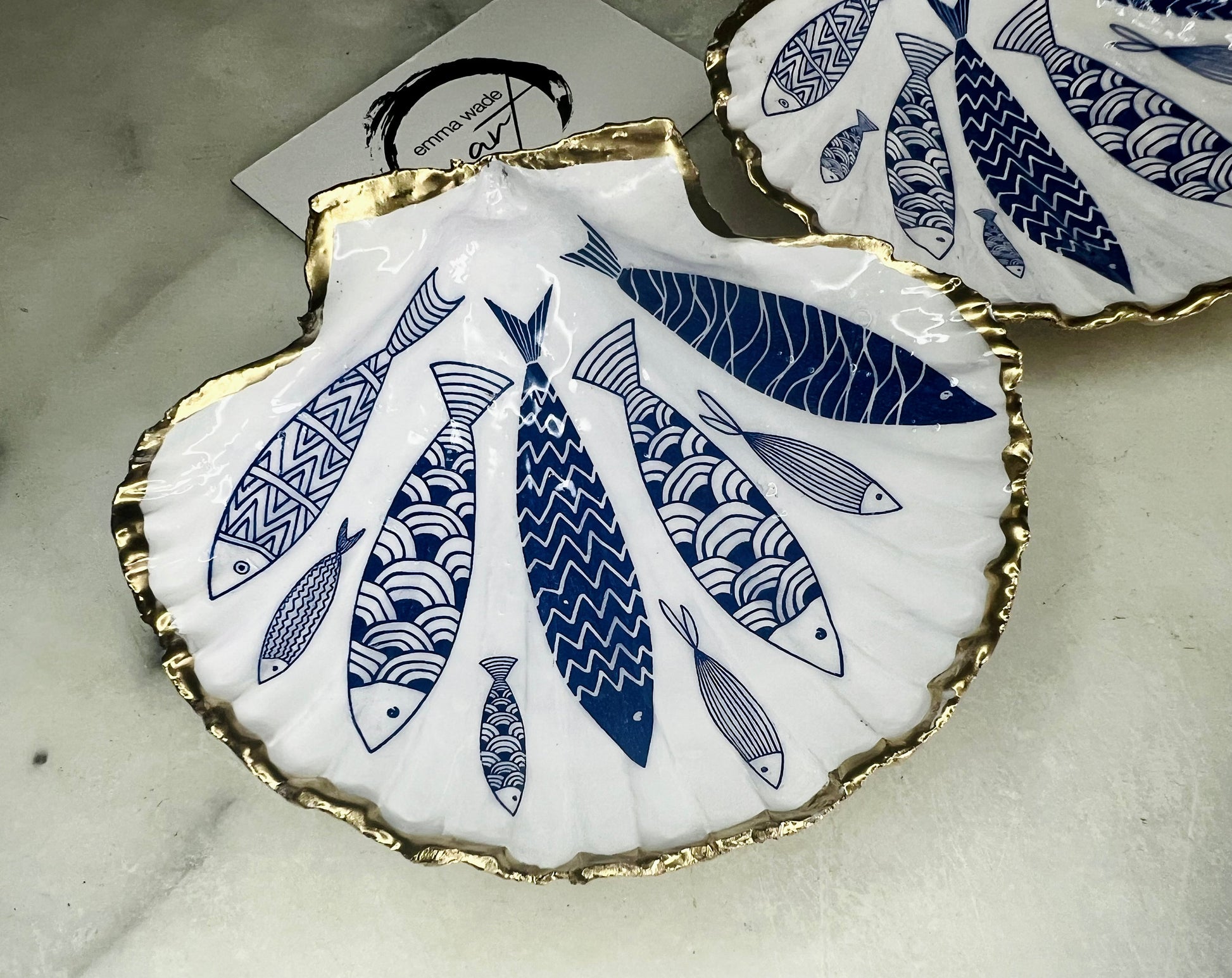 Handmade Dorset scallop shell trinket dishes featuring elegant blue fish illustrations with hand-painted gold leaf edging and glossy crystal clear resin. Coastal nautical jewellery dishes inspired by Scandinavian ocean decor, styled on a neutral background with detailed blue fish artwork and luxurious gilded edges.