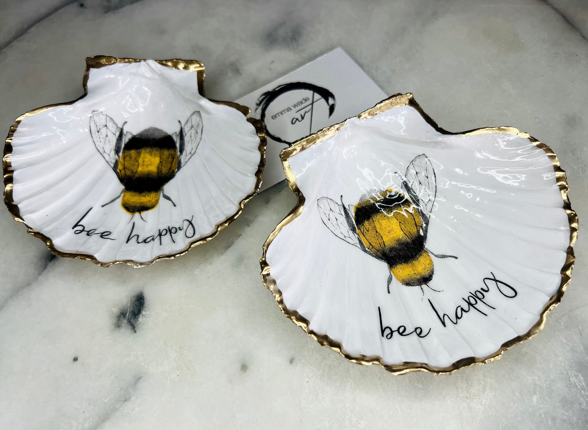 Handmade scallop shell trinket dish featuring a bumblebee illustration with gold leaf edging and glossy resin finish. Coastal jewellery dish in natural white shell with bee artwork, displayed as a ring holder and handmade bee lover gift