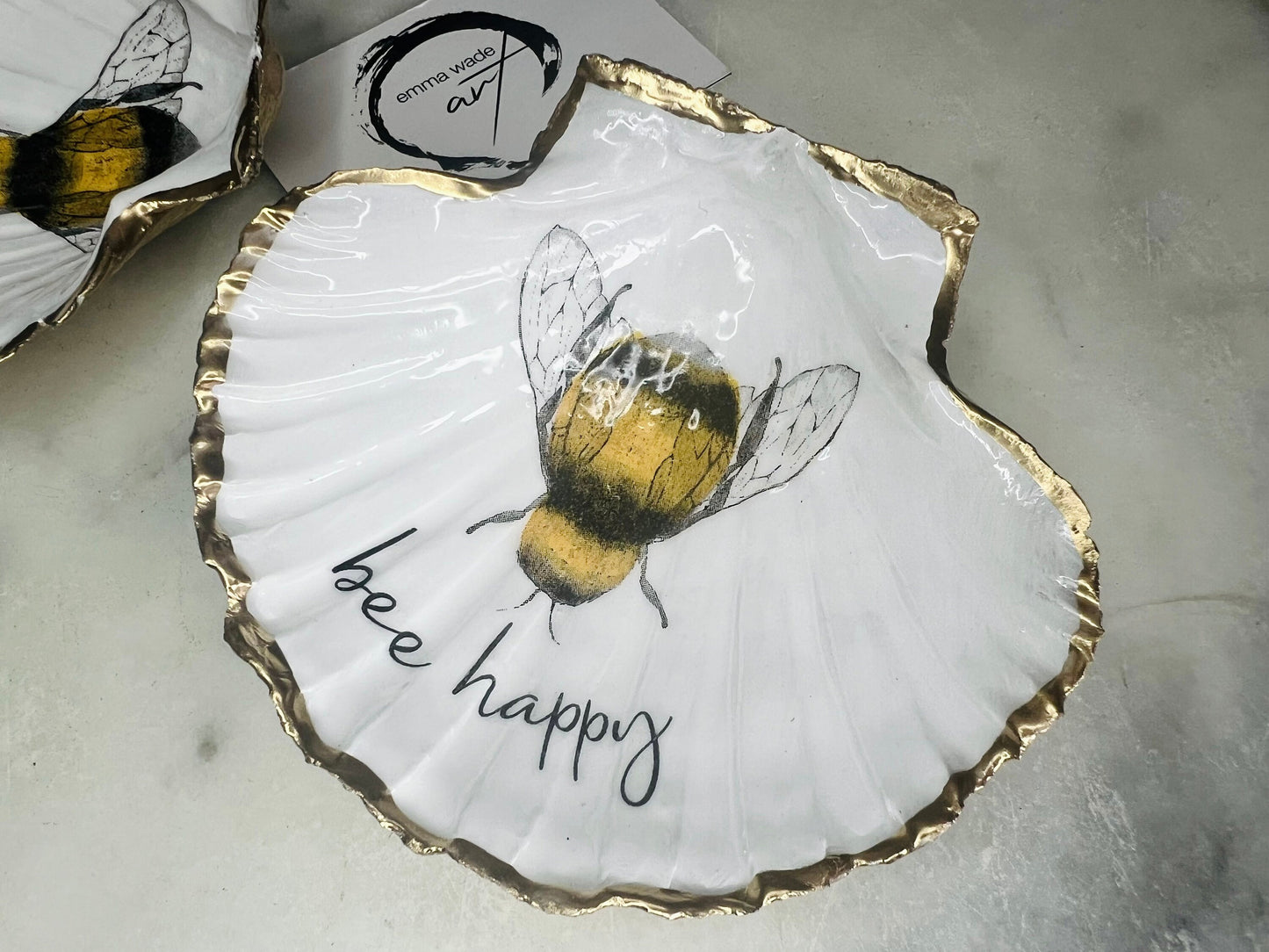Handmade scallop shell trinket dish featuring a bumblebee illustration with gold leaf edging and glossy resin finish. Coastal jewellery dish in natural white shell with bee artwork, displayed as a ring holder and handmade bee lover gift