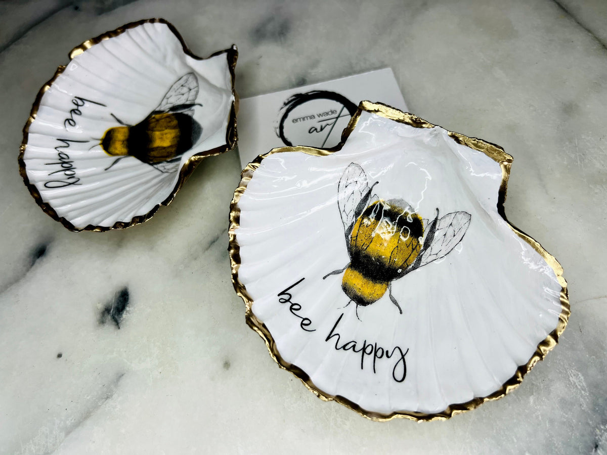 Handmade scallop shell trinket dish featuring a bumblebee illustration with gold leaf edging and glossy resin finish. Coastal jewellery dish in natural white shell with bee artwork, displayed as a ring holder and handmade bee lover gift