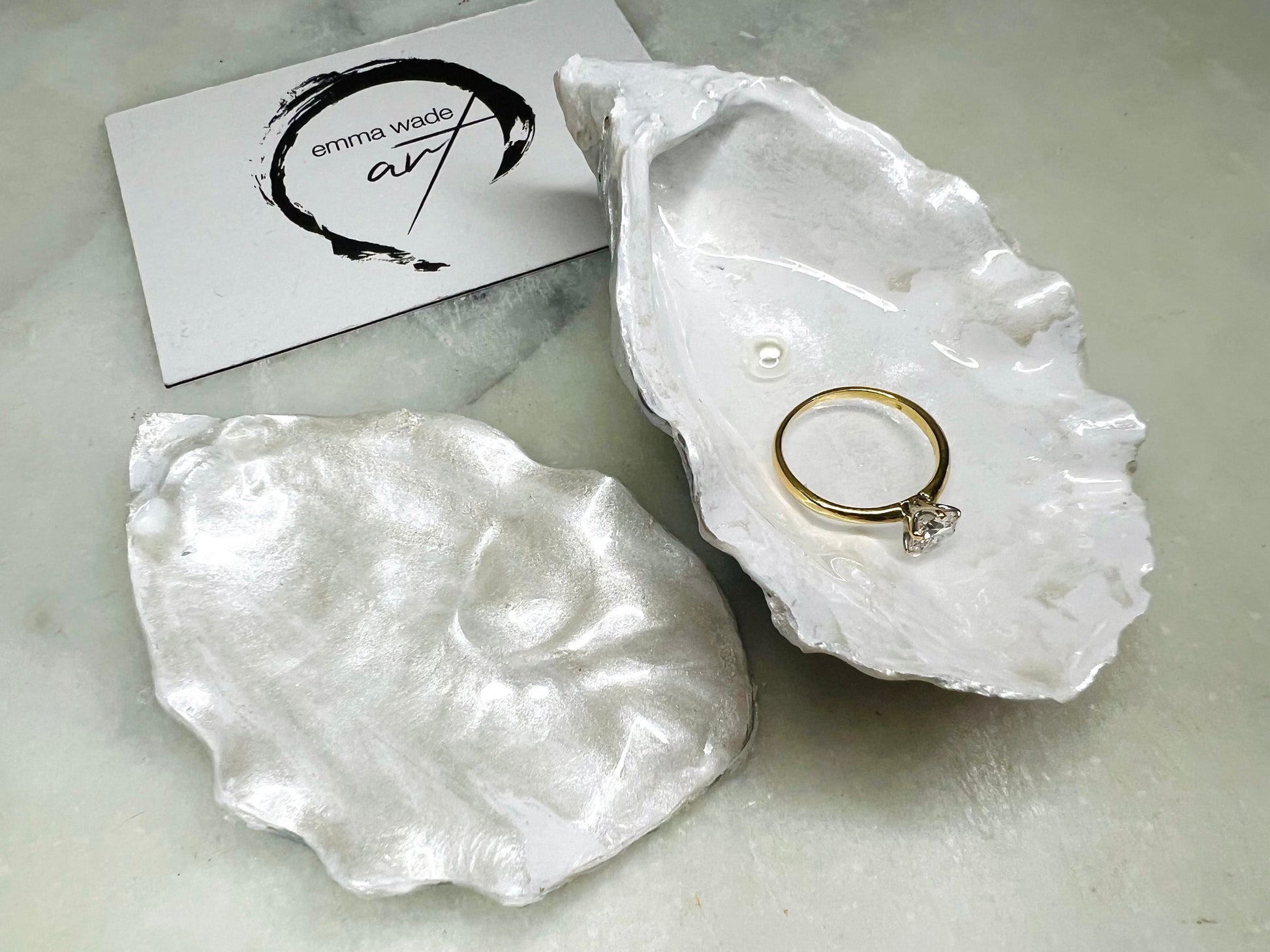 Handcrafted Dorset oyster shell ring box with matching natural shell lid, featuring a luminous inset Swarovski pearl nestled inside the cupped shell. Interior has a soft pearlescent white sheen and is sealed with crystal-clear resin. Optional outer finish in natural pearl tone or pearl with subtle gold leaf highlights. Coastal-inspired engagement proposal ring holder and wedding keepsake, styled on marble surface.