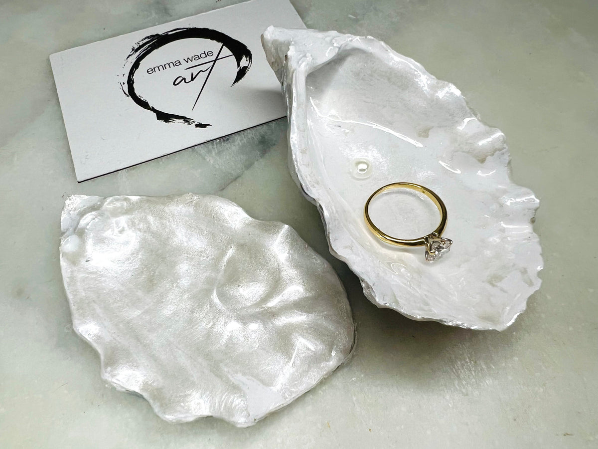 Handcrafted Dorset oyster shell ring box with matching natural shell lid, featuring a luminous inset Swarovski pearl nestled inside the cupped shell. Interior has a soft pearlescent white sheen and is sealed with crystal-clear resin. Optional outer finish in natural pearl tone or pearl with subtle gold leaf highlights. Coastal-inspired engagement proposal ring holder and wedding keepsake, styled on marble surface.